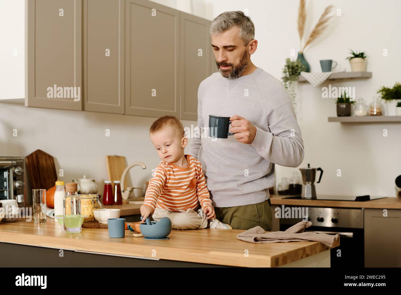 Reifer Vater mit einer Tasse Tee oder Kaffee, der neben seinem kleinen Sohn sitzt und am Küchentisch mit Küchenutensilien beim Frühstück sitzt Stockfoto