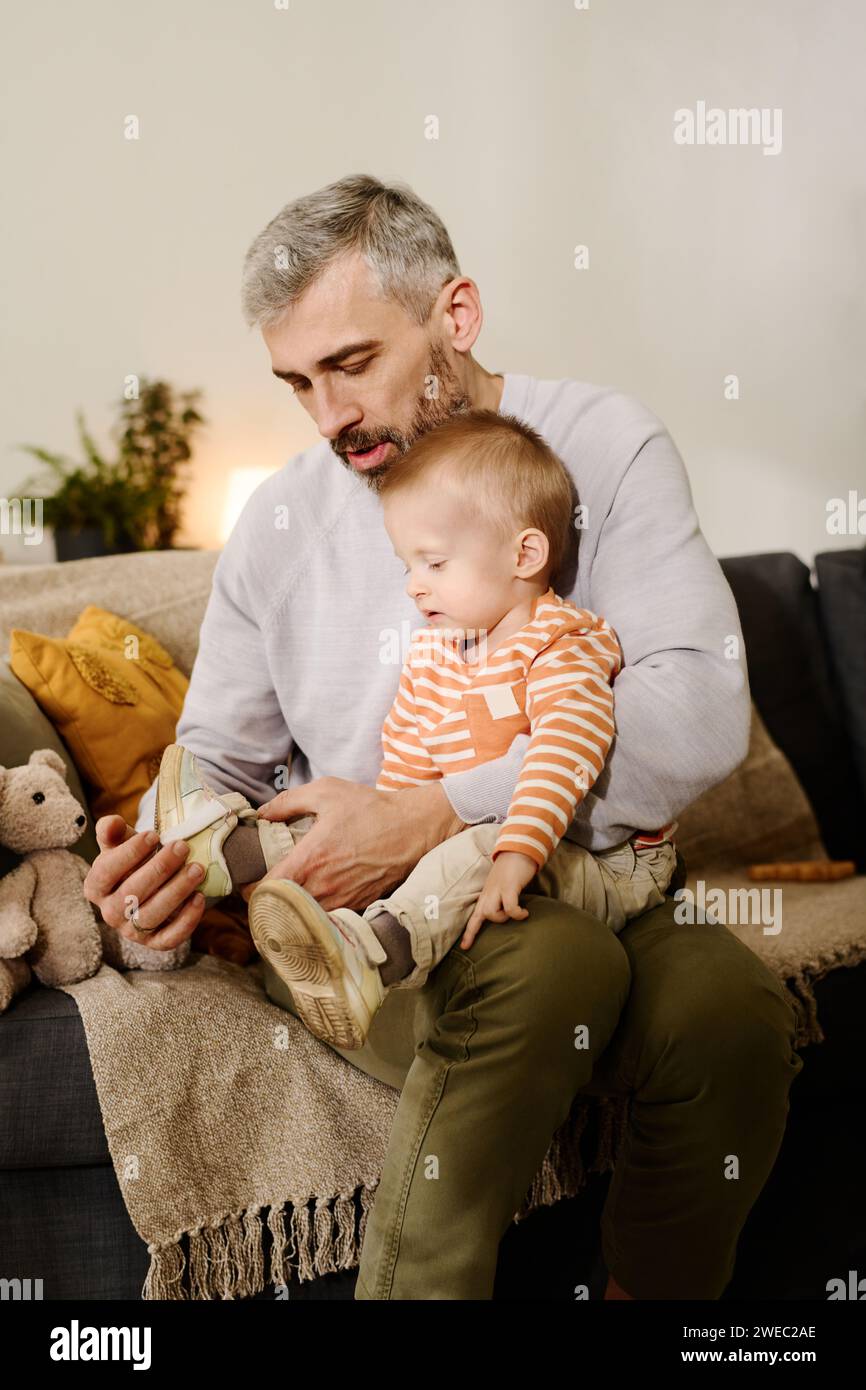 Reifer Vater mit grauen Haaren, der vor der Kamera auf dem Sofa sitzt und den niedlichen Sohn auf den Knien hält, während er Sneakers auf die Füße legt Stockfoto