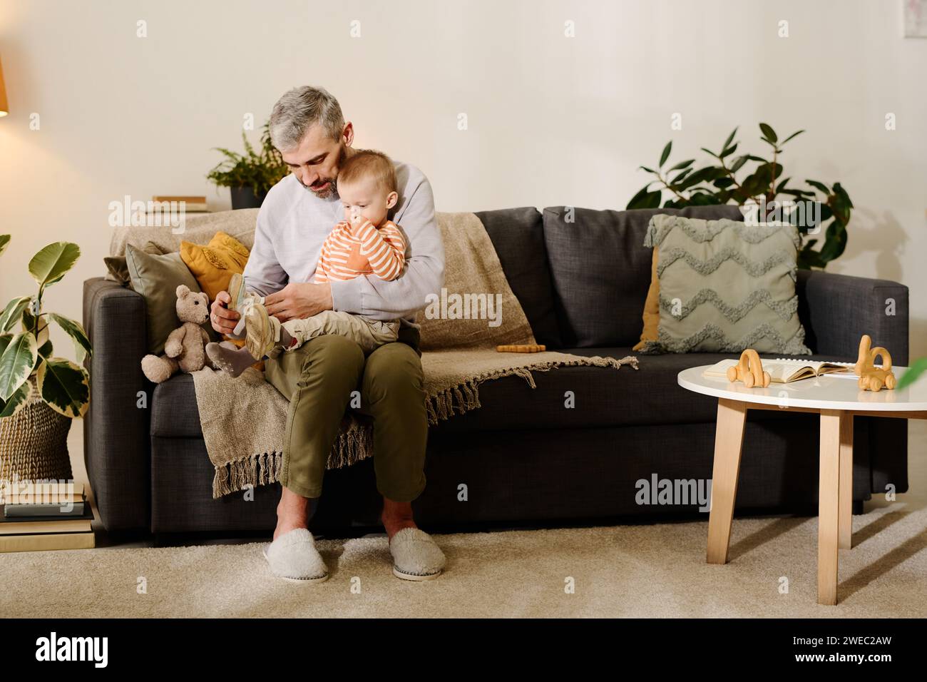 Reifer Mann in Casualwear, der auf der Couch im Wohnzimmer sitzt und den süßen Sohn auf den Knien hält, während er Kreuzschuhe auf die Füße zieht Stockfoto