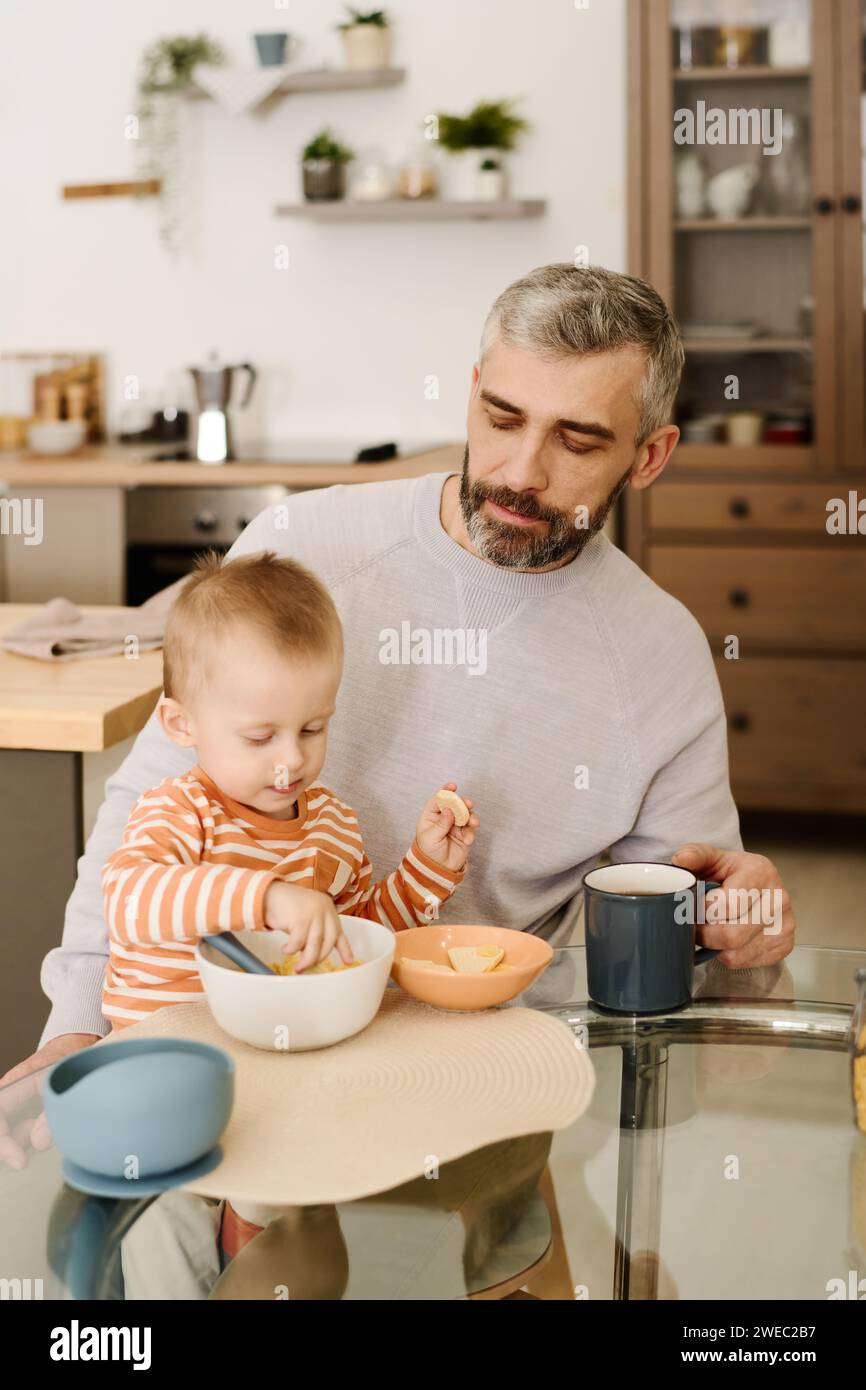 Reifer Mann mit grauen Haaren und Bart, der an einem kleinen runden Glastisch neben seinem niedlichen Sohn sitzt und Kekse zum Frühstück isst Stockfoto