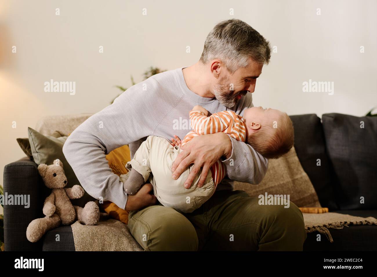 Reifer Mann in Casualwear, der vor der Kamera auf der Couch sitzt und seinen süßen Sohn nach dem Mittagessen einlullt, während er ihn an den Händen hält Stockfoto