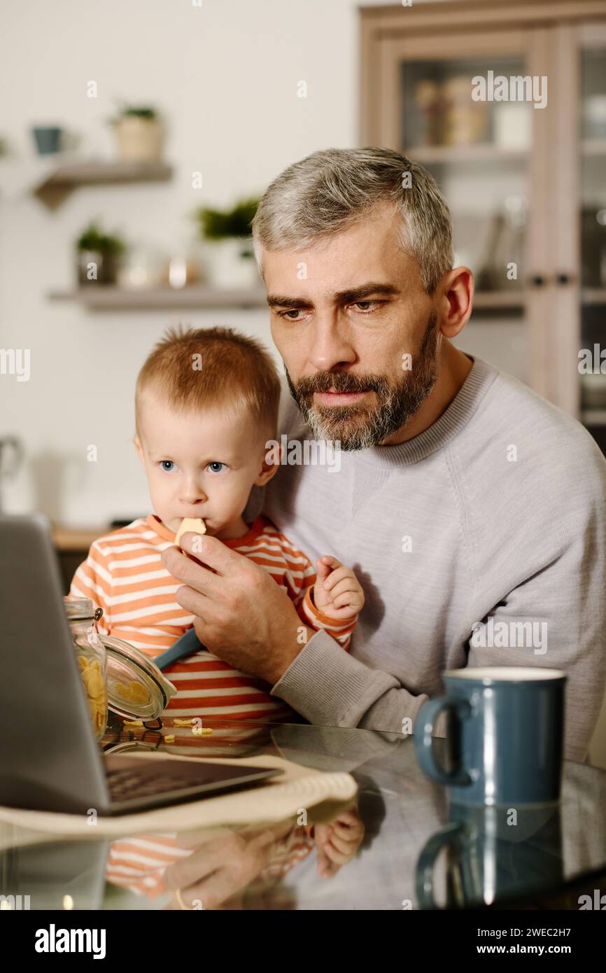 Ernsthafter reifer Mann, der beim Frühstück auf den Laptop-Bildschirm schaut, während er am Küchentisch sitzt und seinen kleinen Sohn mit Cornflakes füttert Stockfoto