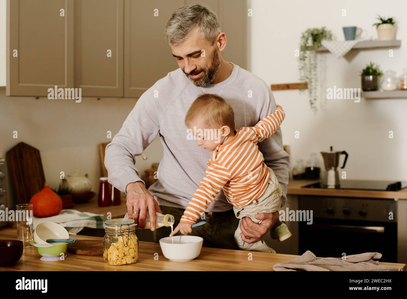 Reifer Mann mit süßem Baby-Sohn auf Händen, der frische Milch in eine Schüssel mit Cornflakes gießt, während er das Frühstück an der Küchentheke zubereitete Stockfoto