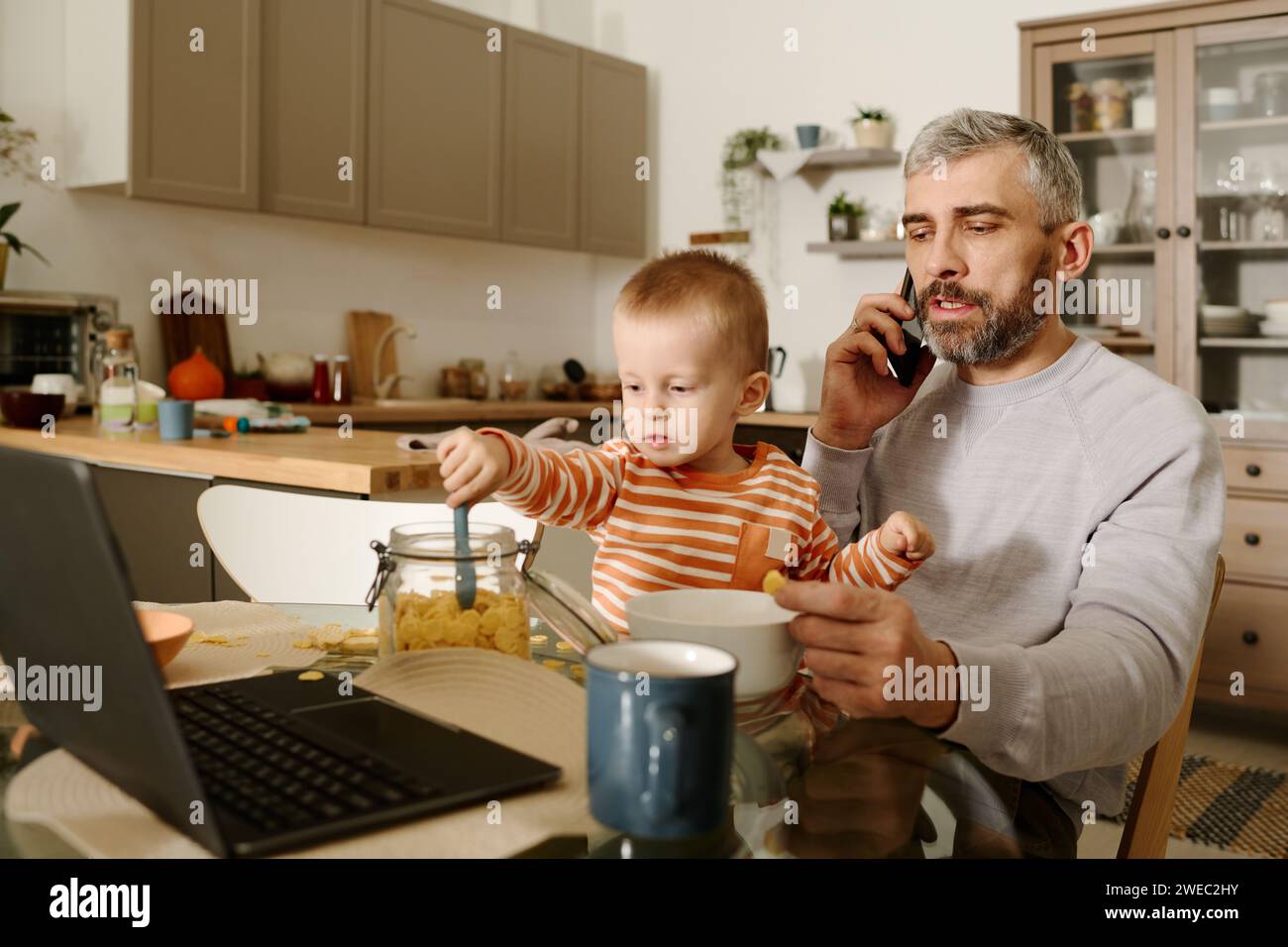 Selbstbewusster, reifer Geschäftsmann mit einem kleinen Sohn auf den Knien, der mit einem Kollegen oder einem Untergebenen auf dem Smartphone am Küchentisch spricht Stockfoto