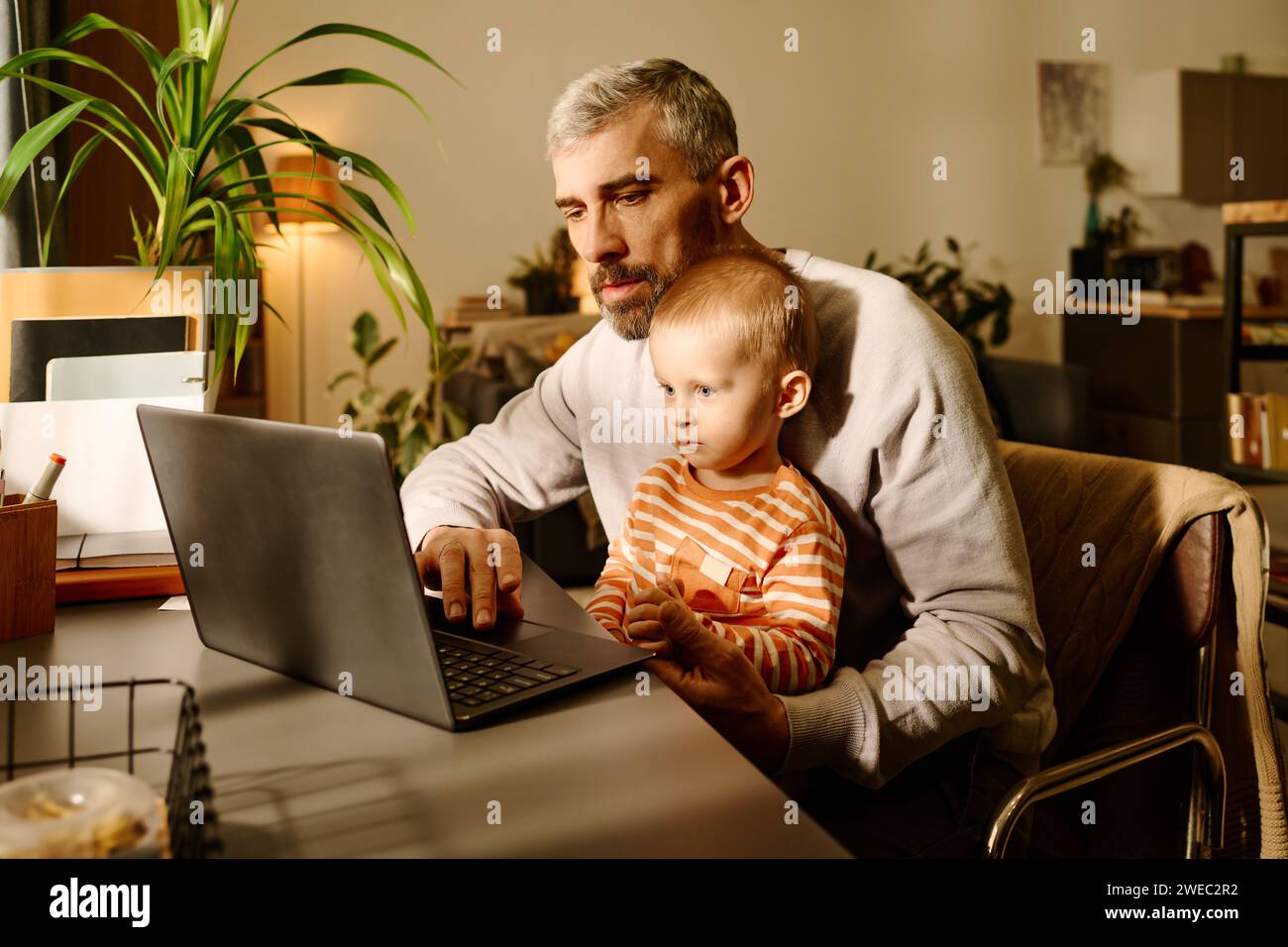 Reifer Home Office Manager oder Solopreneur mit seinem kleinen Sohn, der während des Netzwerks oder beim Ansehen von Online-Videos auf den Laptop-Bildschirm schaut Stockfoto