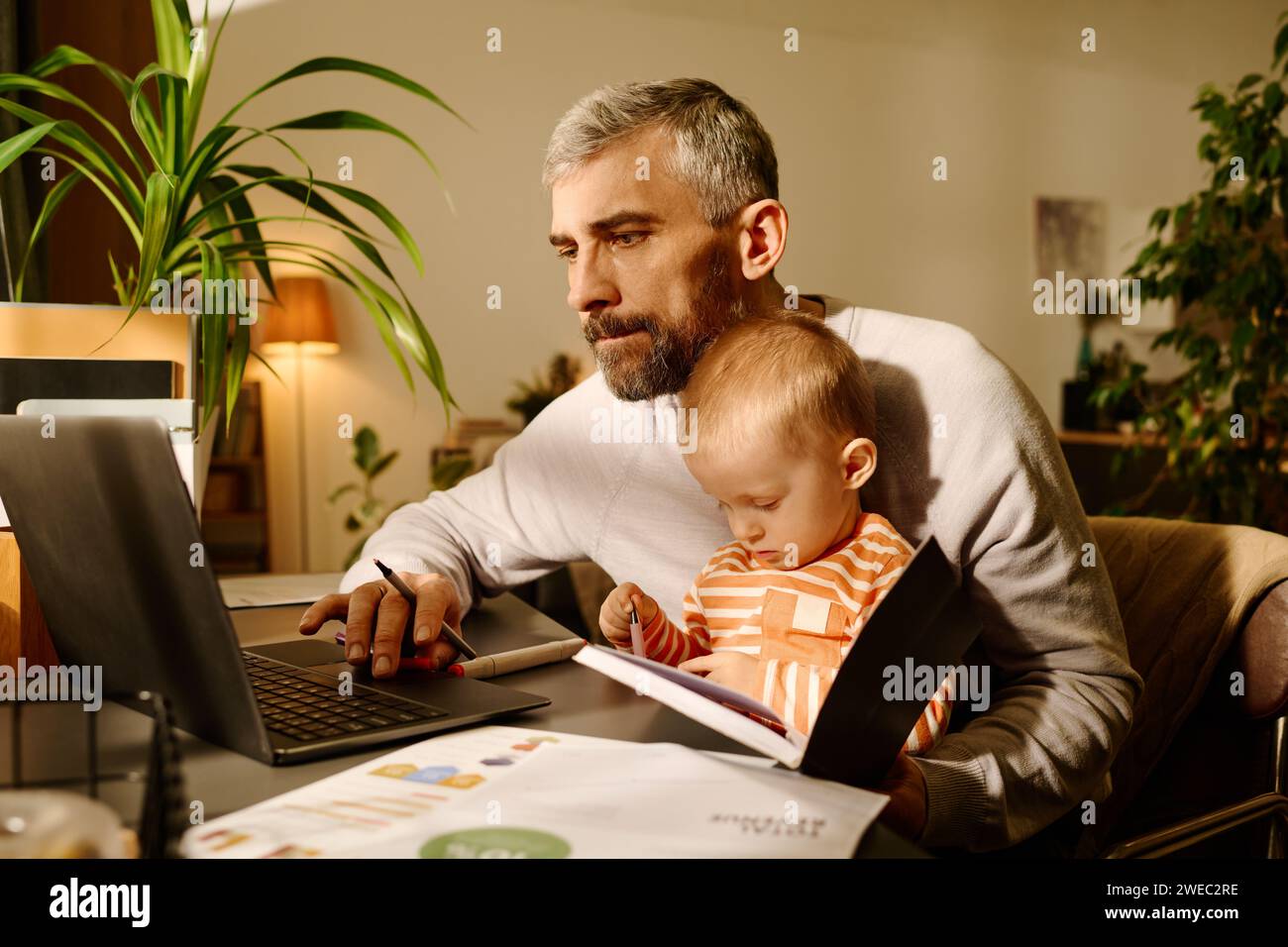Geschäftsmann in Casualwear, der auf den Laptop-Bildschirm blickt, während er am Tisch sitzt und seinen süßen Sohn auf den Knien hält und auf die Seite des offenen Buches blickt Stockfoto