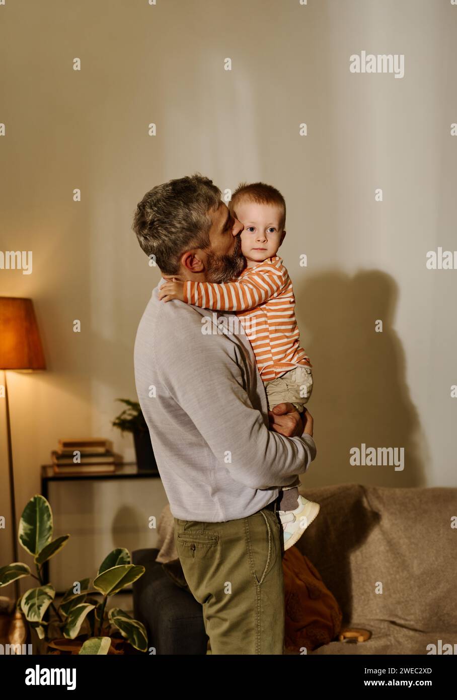 Reifer Vater in Casualwear hält seinen niedlichen Sohn an den Händen und küsst ihn auf die Wange, während er zu Hause vor der Kamera steht Stockfoto