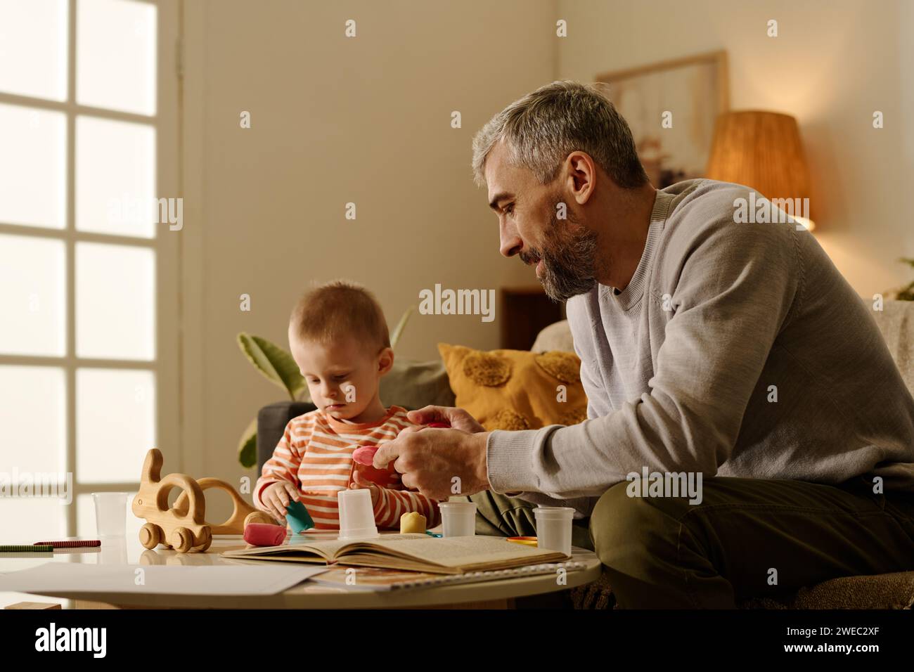 Reifer Mann im grauen Pullover, der neben dem entzückenden Baby-Sohn an einem kleinen runden Tisch sitzt und mit ihm in der Freizeit mit neuen Spielzeugen spielt Stockfoto