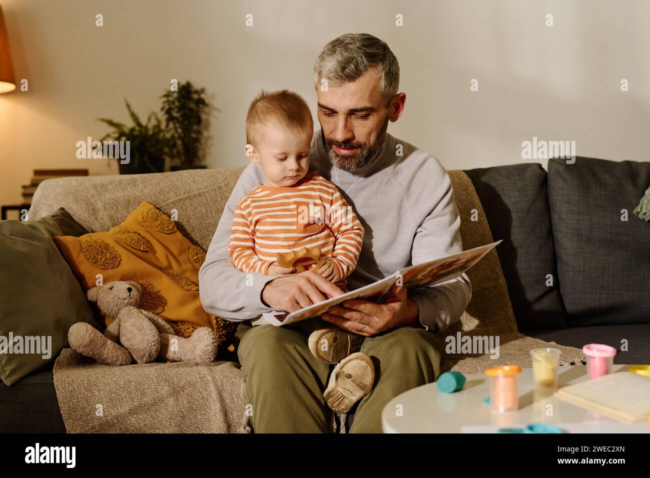 Reifer Vater in Casualwear sitzt auf der Couch mit seinem niedlichen Sohn auf den Knien und zeigt auf die Seite des Buches, während er ein Märchen liest Stockfoto