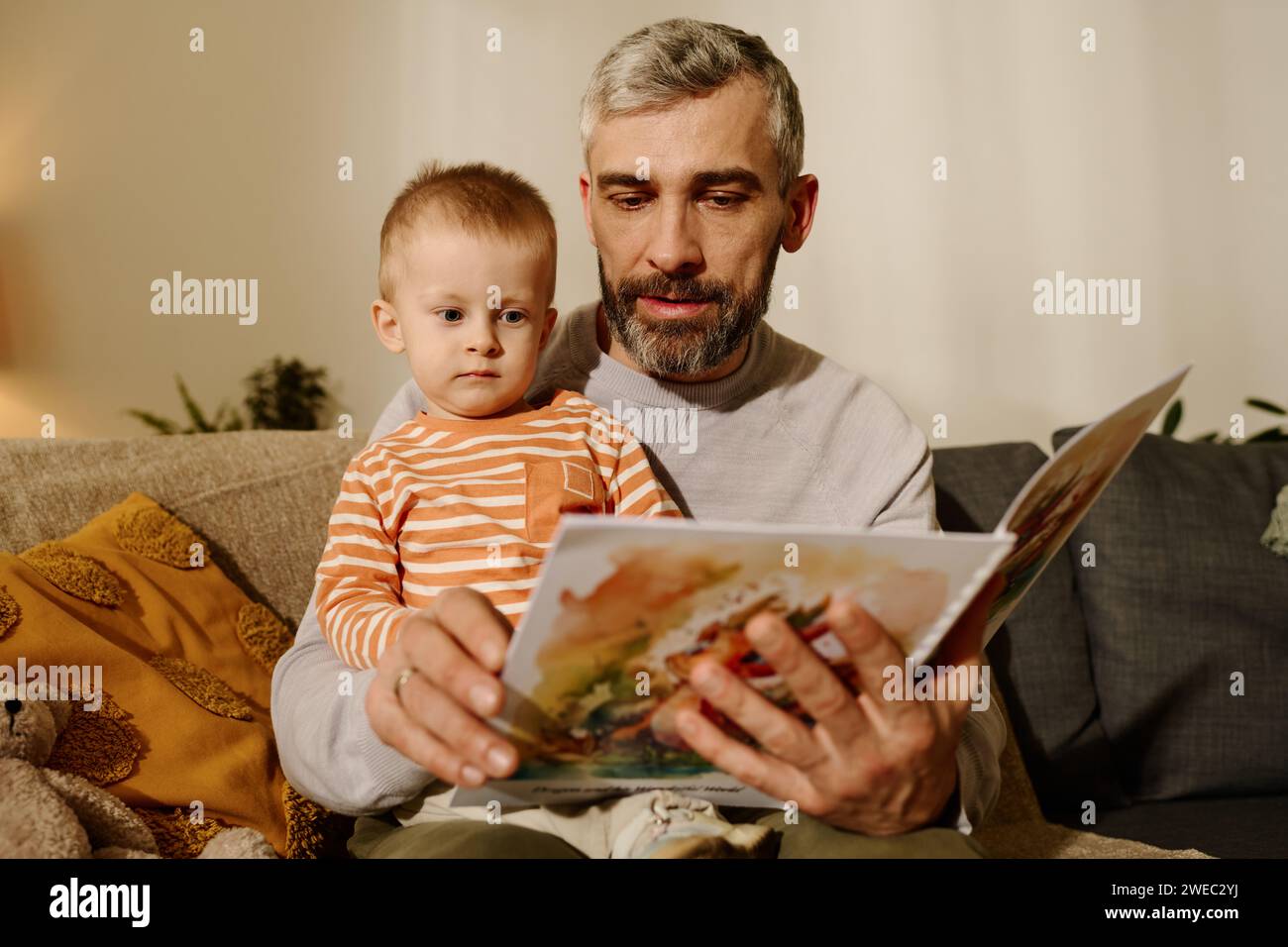 Entzückender Junge, der sich die Seite eines offenen Buches mit Comics oder Märchen anschaut, während er auf den Knien seines Vaters sitzt, der ihm die Geschichte vorliest Stockfoto