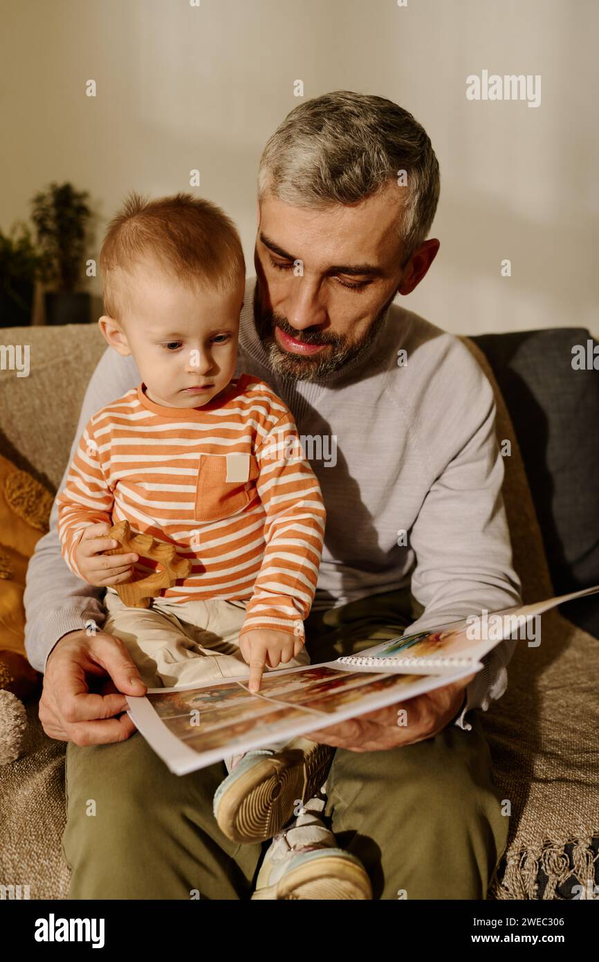 Reifer Vater mit offenem Comicbuch und entzückendem Sohn, der auf eine Seite zeigt, mit Bild, das vor der Kamera auf der Couch sitzt Stockfoto