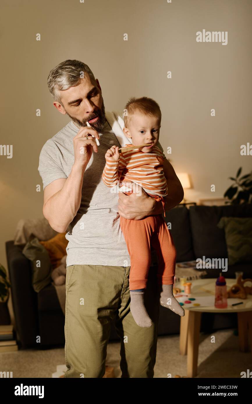 Reifer Mann in T-Shirt und Hose, der Zähne putzt und den niedlichen Sohn an seinen Händen ansieht, während er vor der Kamera steht Stockfoto