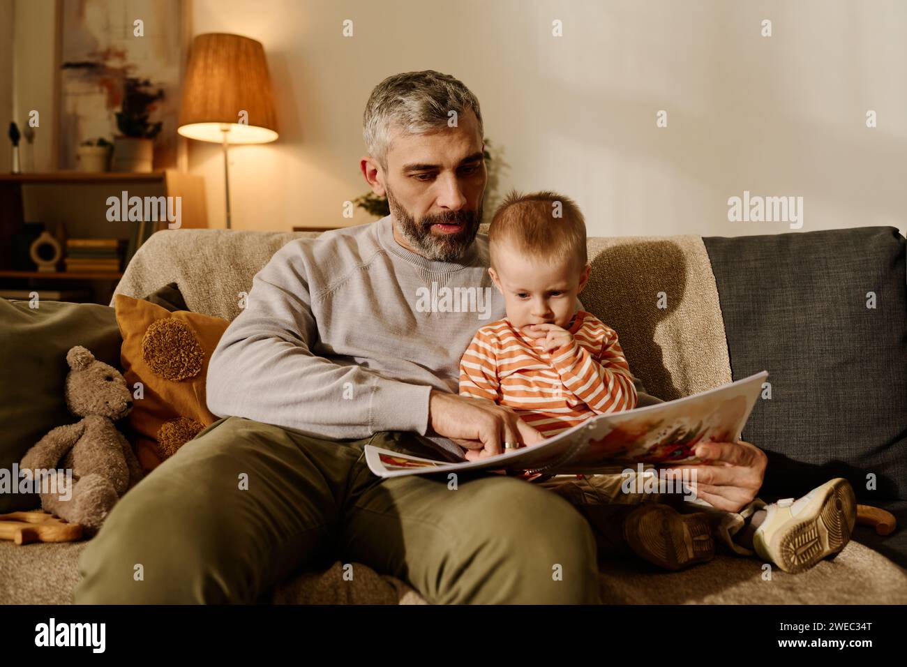 Reifer Vater zeigt auf ein Bild auf der Seite eines offenen Buches mit Comics oder Märchen, während er es seinem liebenswerten kleinen Sohn in Ruhe vorliest Stockfoto