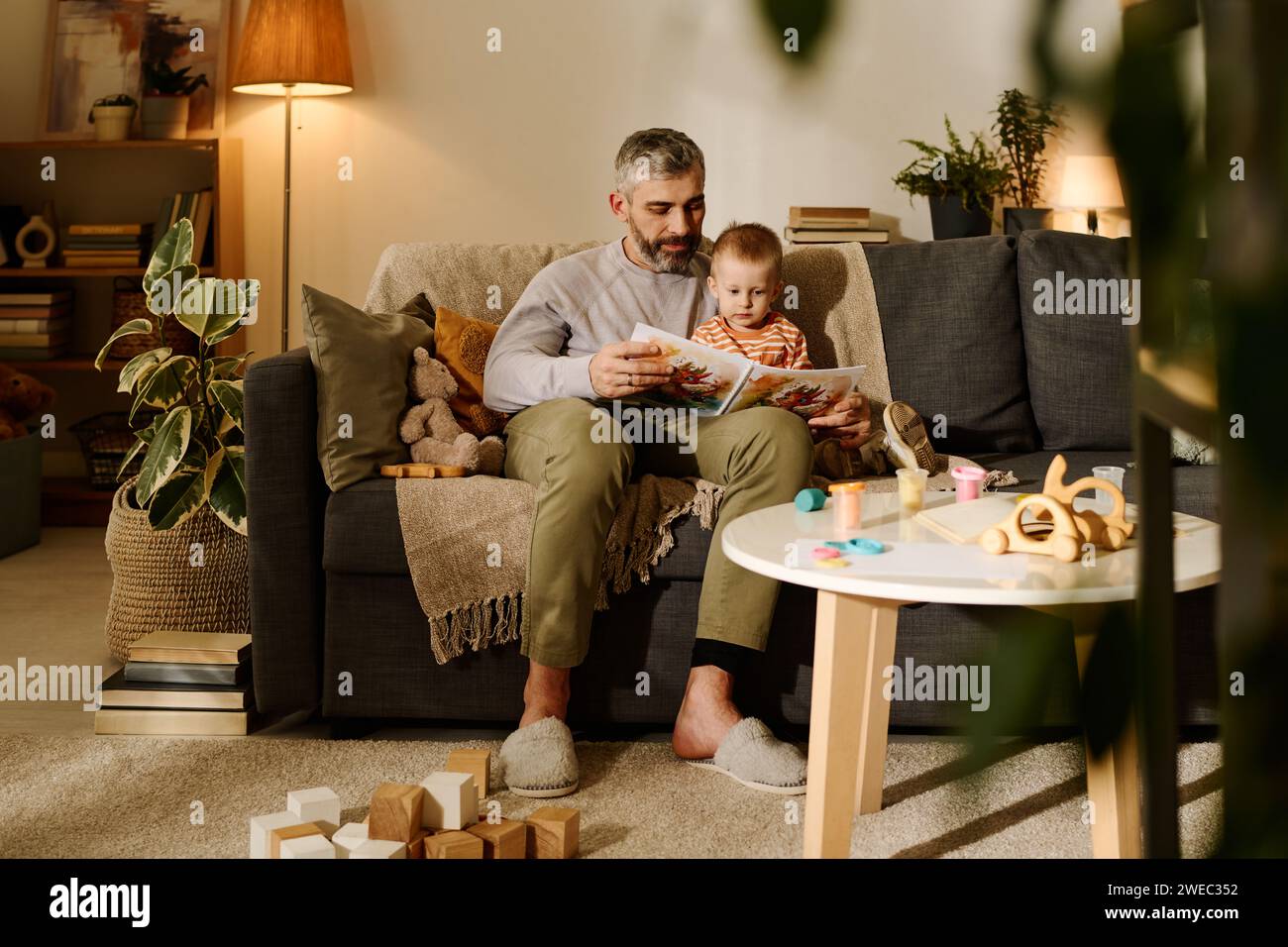 Reifer Vater liest seinem süßen Sohn Märchenbuch vor, während er auf der Couch vor einem kleinen runden Tisch im Wohnzimmer sitzt Stockfoto