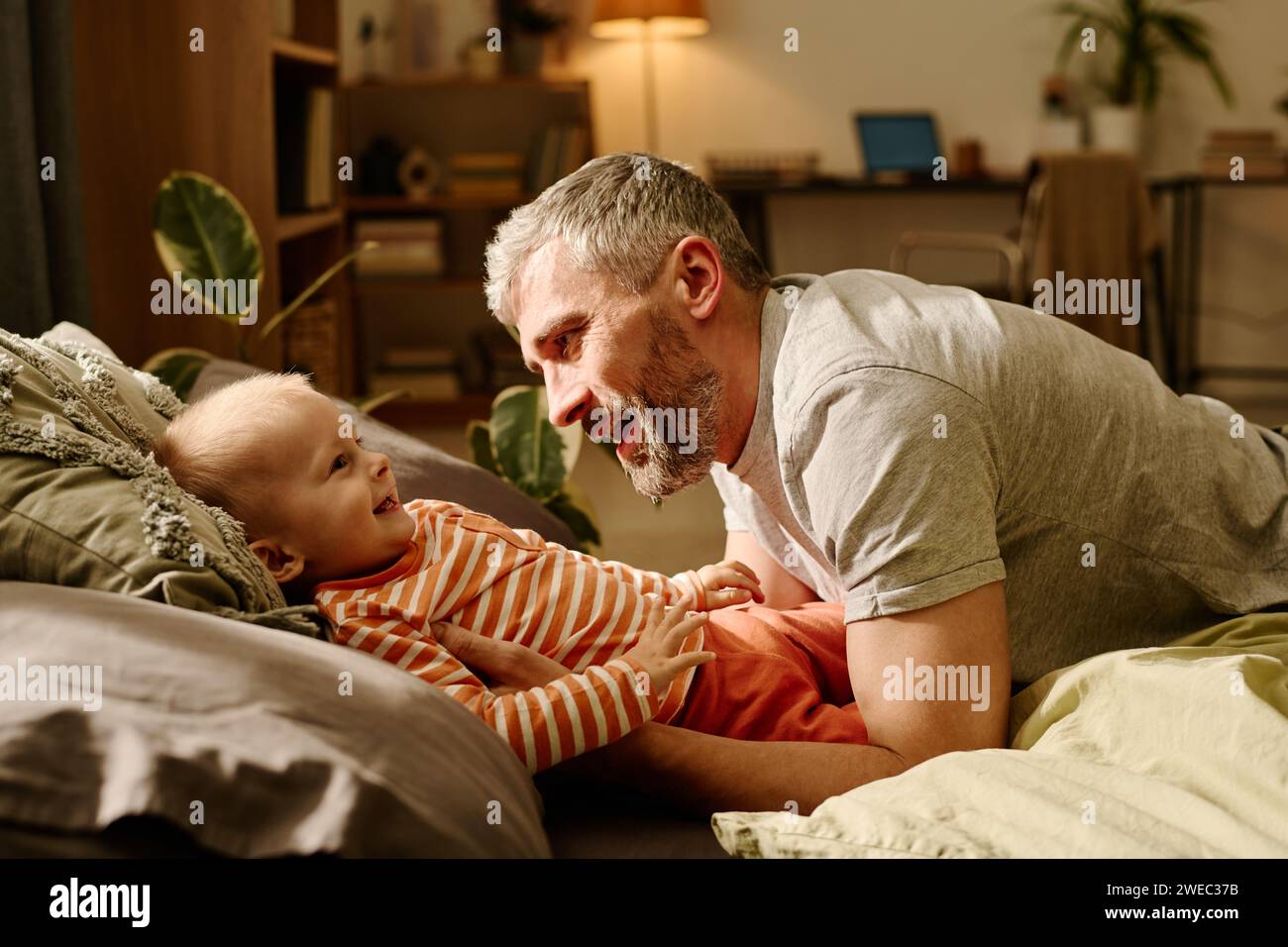 Seitenansicht eines glücklichen reifen Mannes mit grauen Haaren und Bart, der mit seinem süßen Sohn auf dem Bett oder der Couch im Wohnzimmer spielt Stockfoto