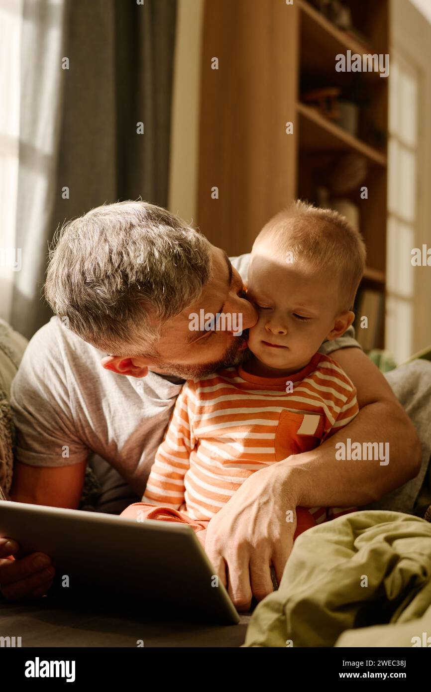 Liebevoller Vater, der seinen entzückenden Sohn auf die Wange küsst und ihn umarmen kann, während er sich in der Freizeit entspannt und Online-Videos ansieht Stockfoto