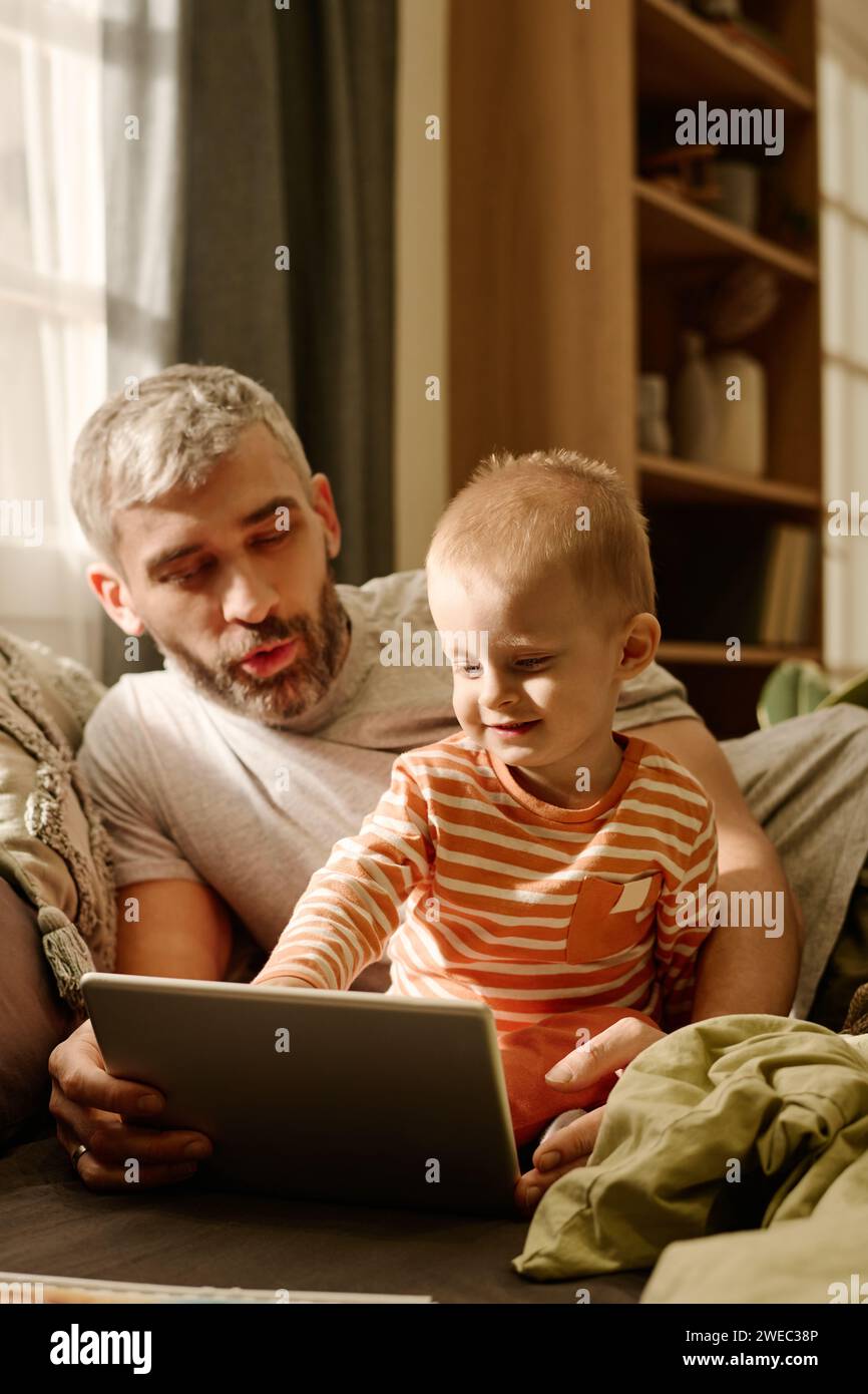 Reifer Vater und kleiner Sohn schauen auf den Tablet-Bildschirm, während sie sich auf dem Bett oder der Couch vor der Kamera entspannen und gemeinsam Online-Videos ansehen Stockfoto