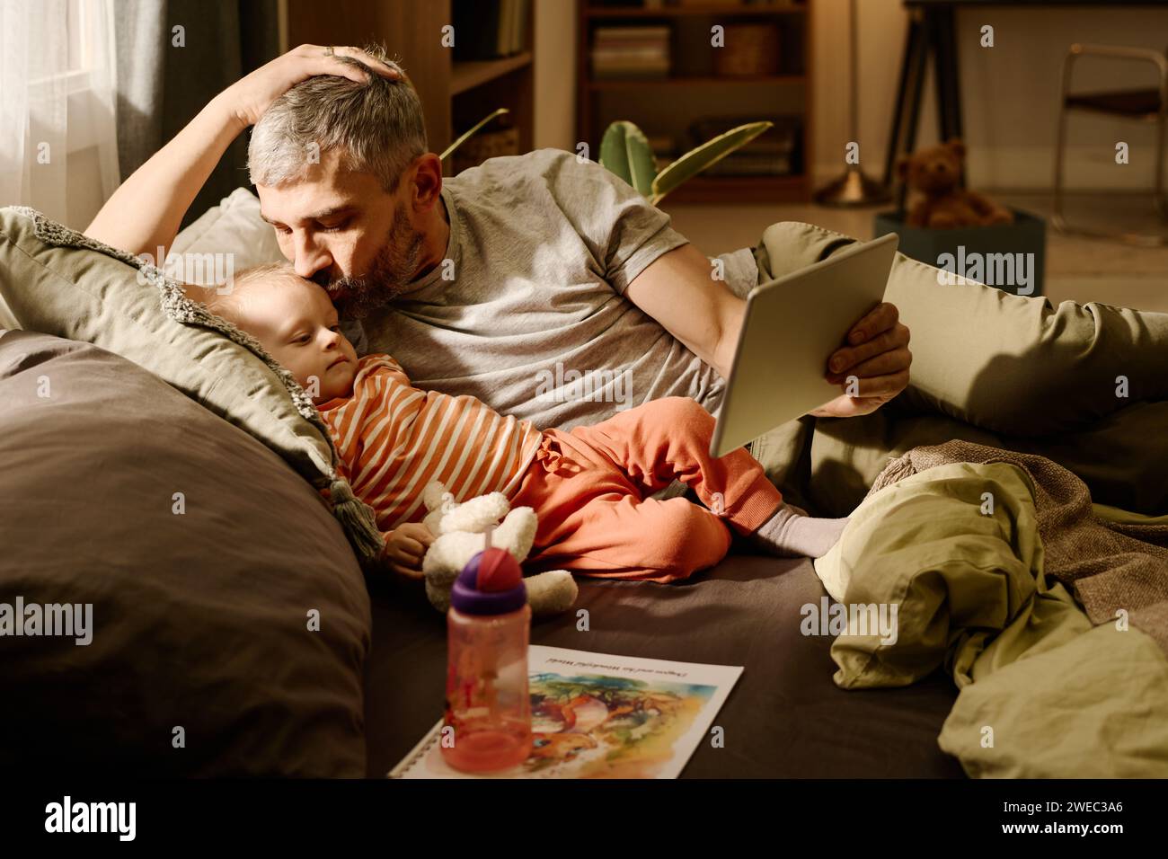 Mann mit grauen Haaren, der seinen niedlichen Sohn küsst, der neben ihm liegt und auf den Bildschirm eines Tablets blickt, der von reifem Vater gehalten wird Stockfoto