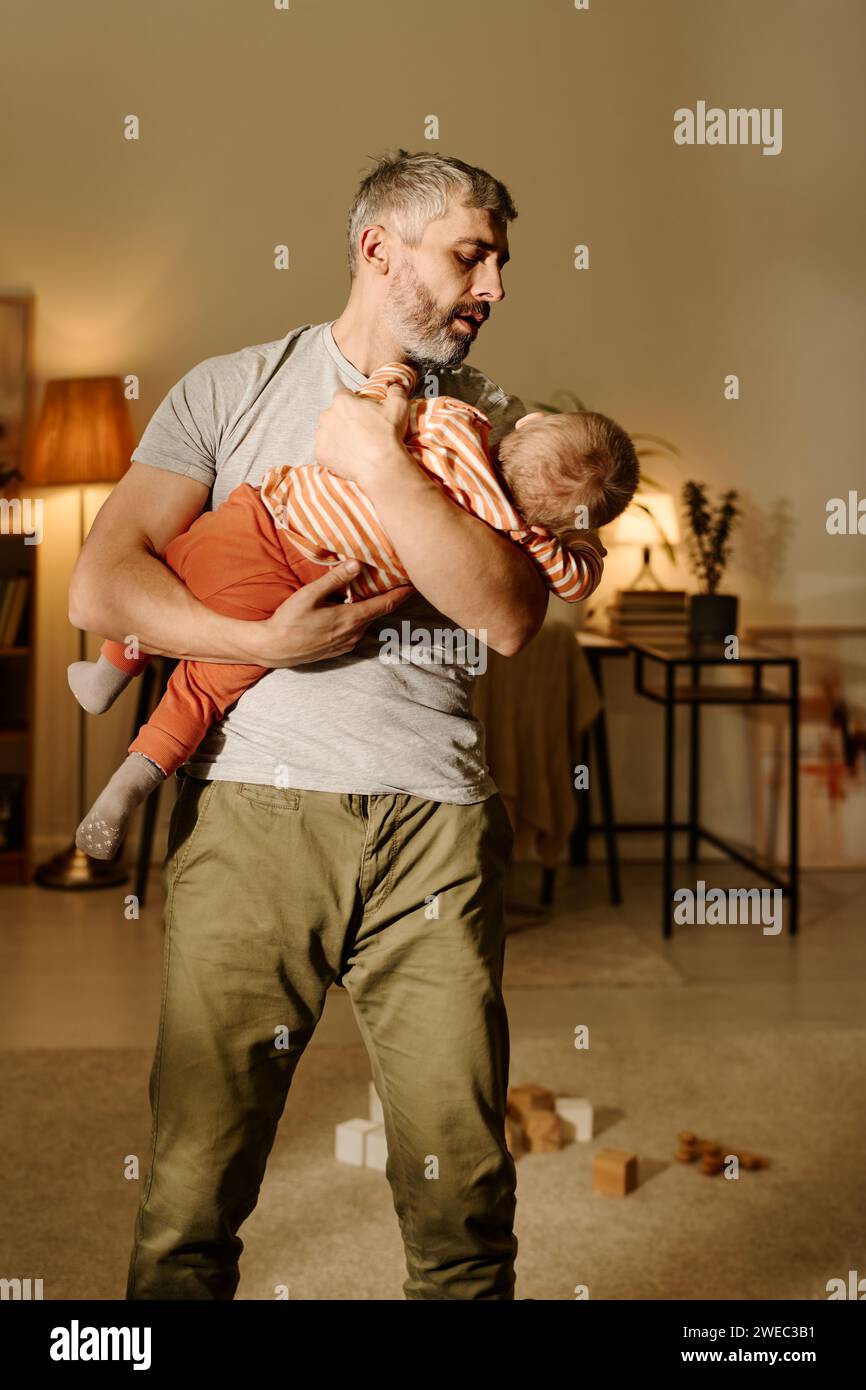 Reifer Vater in Casualwear hält seinen niedlichen Sohn an den Händen und sieht ihn an, während er im Wohnzimmer vor der Kamera steht Stockfoto