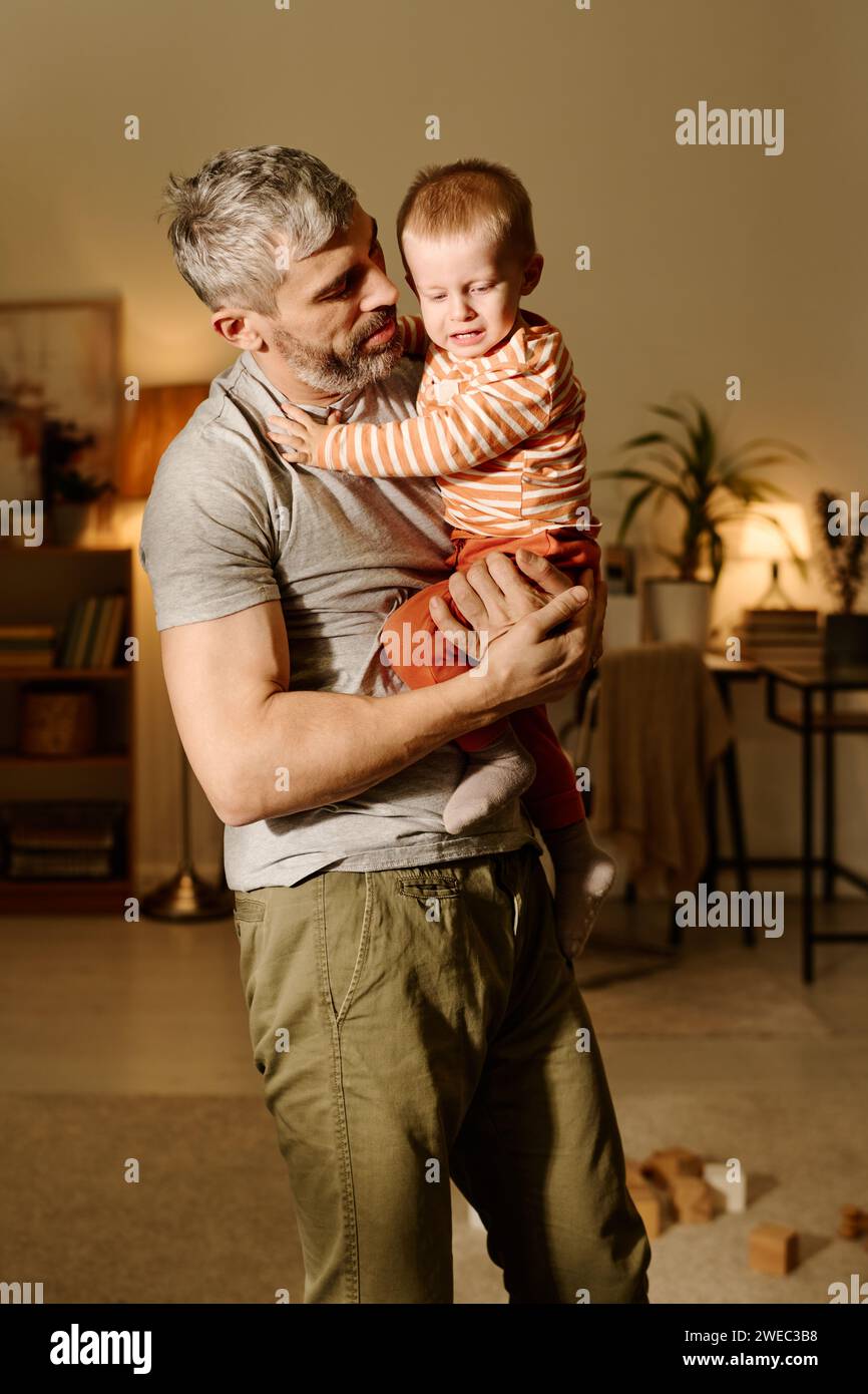 Reifer Mann in grauem T-Shirt und beigefarbener Hose, der einen schreienden, frechen Sohn an den Händen hält und ihn ansieht, während er im Wohnzimmer steht Stockfoto