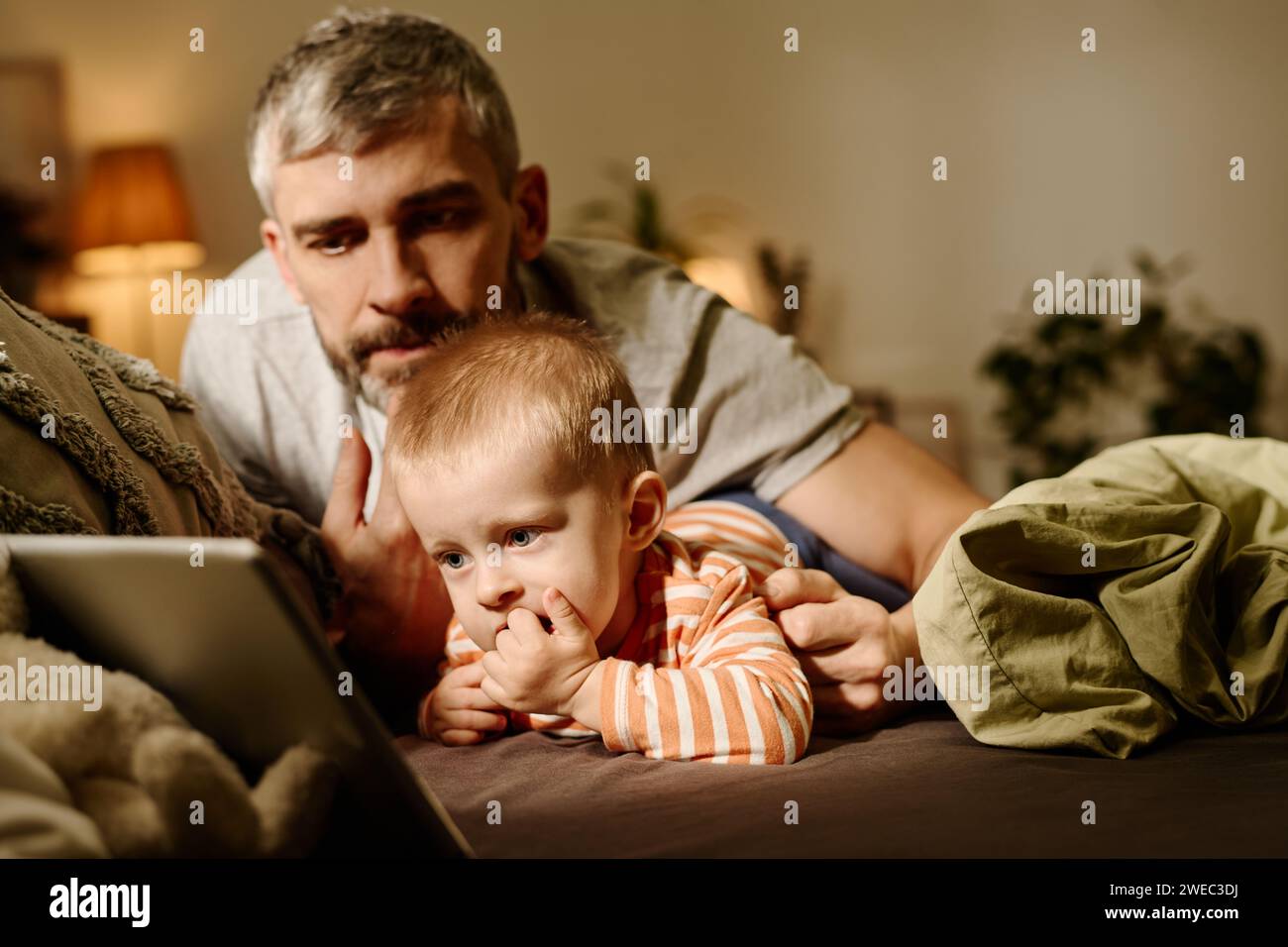 Reifer Vater liegt neben dem süßen Baby Sohn auf dem Bett, während er auf den Tablet-Bildschirm blickt und am Abend Online-Cartoons ansieht Stockfoto