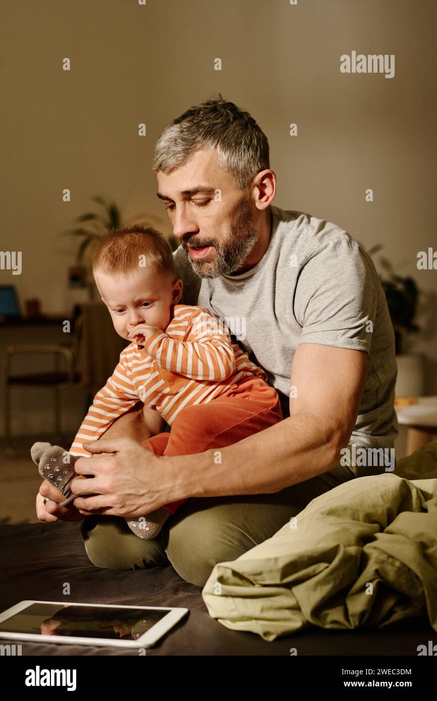Reifer Mann, der kleine graue Socken auf die Füße seines liebenswerten kleinen Sohnes legt, der auf den Knien seines Vaters sitzt und auf den Tablet-Bildschirm blickt Stockfoto
