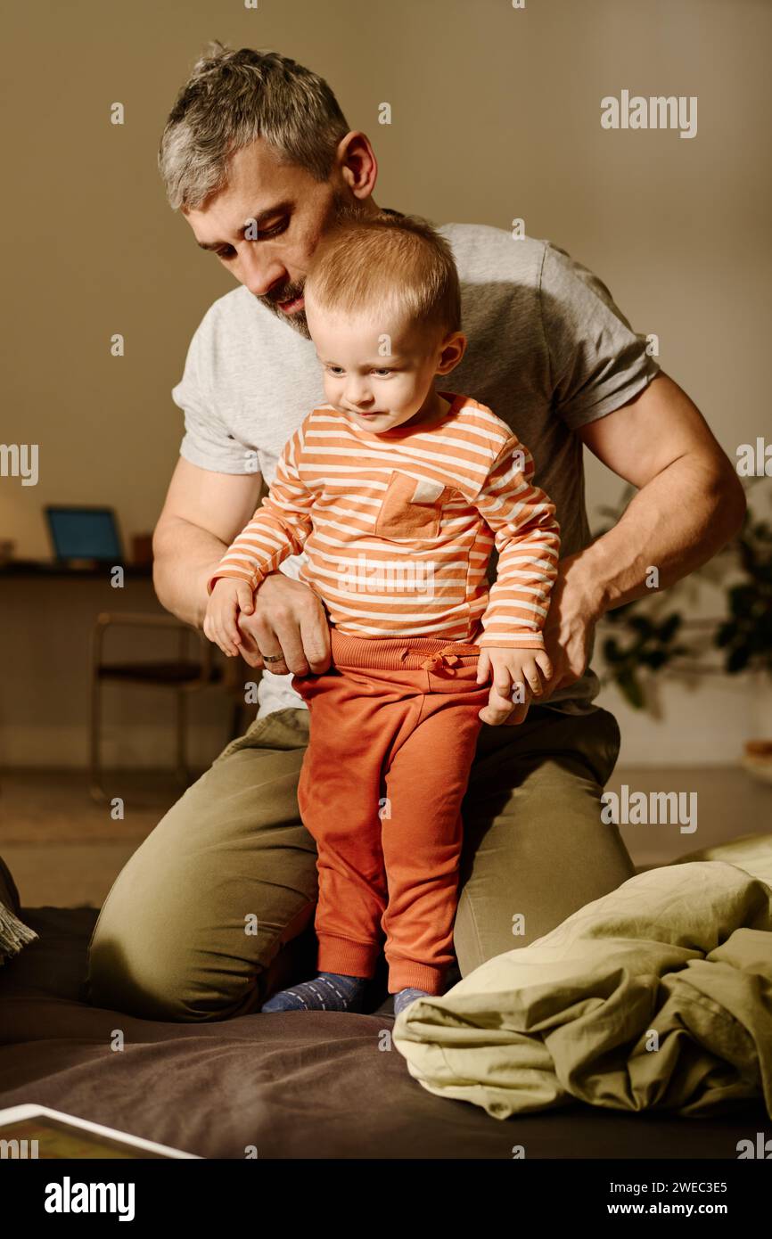 Der reife Vater zieht seinem niedlichen Sohn eine Hose an, der neben ihm auf dem Bett steht und sich ein Online-Video auf dem Tablet-Bildschirm ansieht Stockfoto