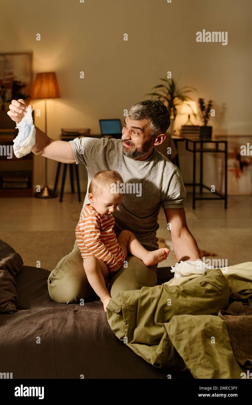 Ein ekelhafter Vater, der die schmutzige Windel seines kleinen Sohnes ansah, während er auf dem Bett saß, mit einem entzückenden Jungen auf den Knien und einer Windel Stockfoto
