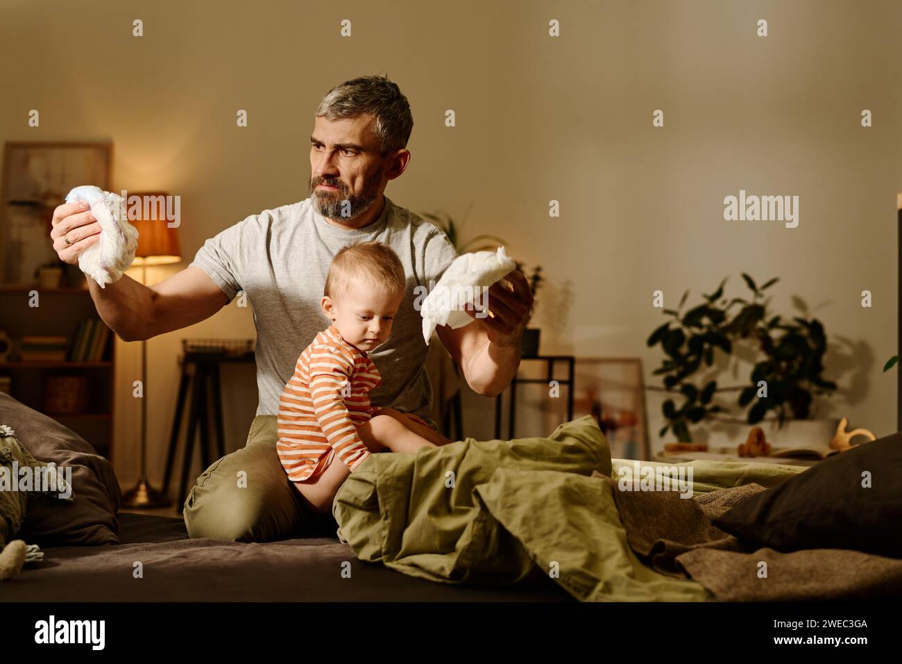 Reifer, angeekelter Mann, der schmutzige Windel in der Hand ansieht, während er auf dem Bett sitzt, mit einem niedlichen Sohn auf den Knien und Windeln wechselt Stockfoto