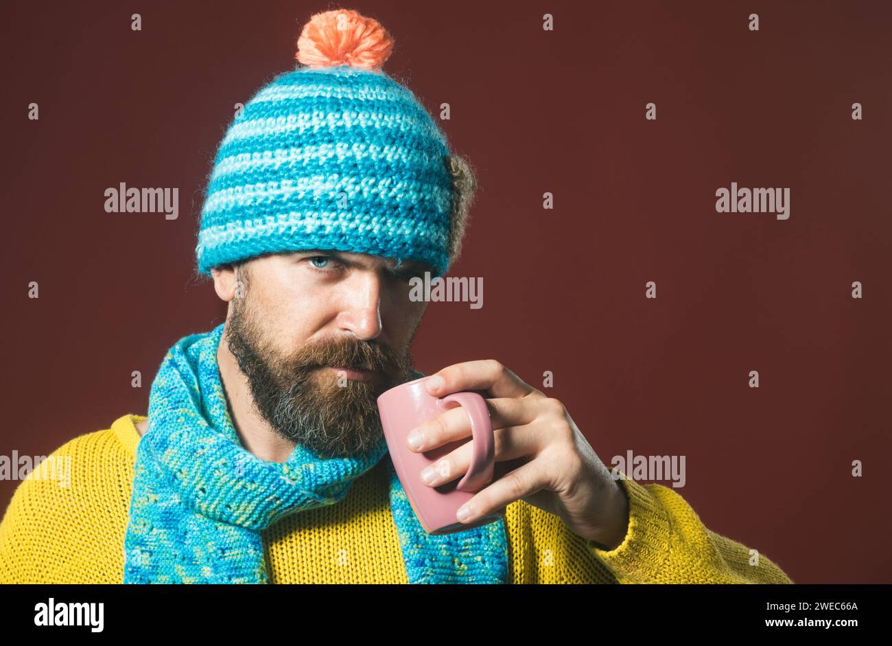 Ernster Mann mit Bart und Schnurrbart in gelbem Pullover, der eine Tasse heißen Kaffee oder Tee trinkt. Herbst- oder Winterzeit. Bärtiger Mann mit Hut und Schal trinken Stockfoto