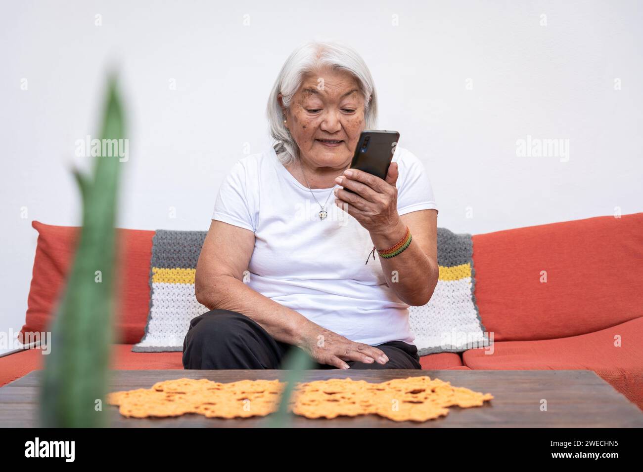Ältere Frau japanischer Herkunft mit grauen Haaren, die auf den Bildschirm ihres Mobiltelefons blickt und zu Hause sitzt. Online-Kommunikation über ein Smartphone. Stockfoto
