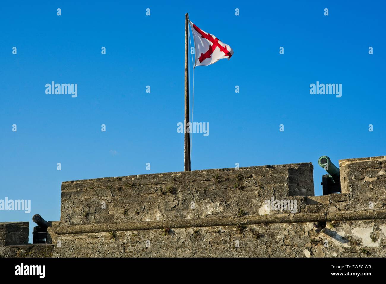 Spanisches Kolonialkreuz von Burgund Flagge über Reihe von Kanonen auf der Coquina-Steinmauer der Festung Castillo de San Marcos in St. Augustine Florida Stockfoto