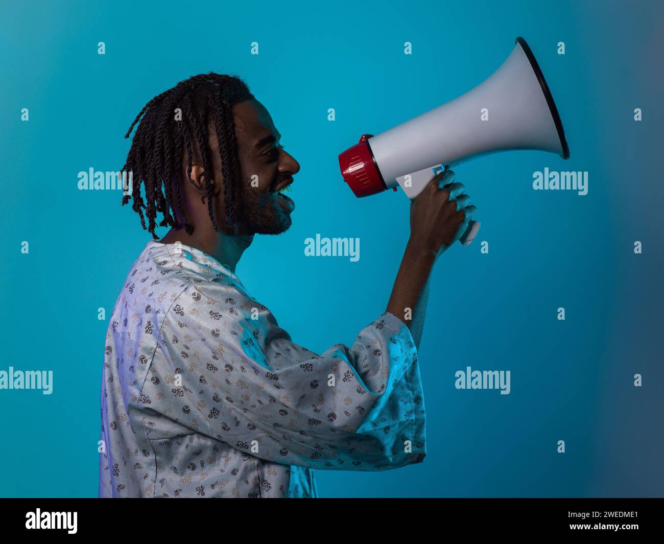Der afroamerikanische Mann trägt traditionelle Kleidung und verwendet leidenschaftlich ein Megaphon vor einem auffälligen blauen Hintergrund, das sein Gesangsbild symbolisiert Stockfoto