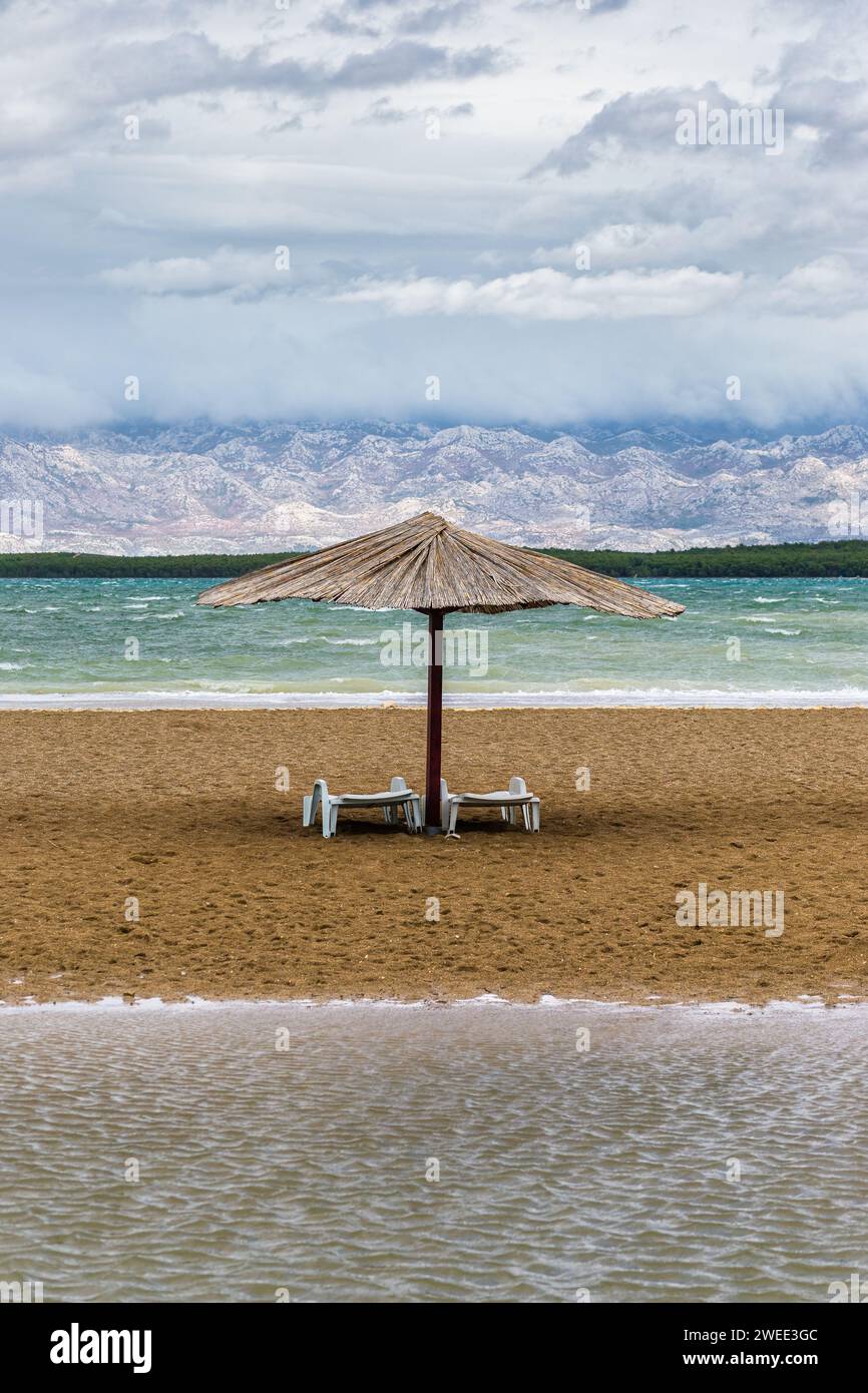 Nin, Kroatien - Schilfsonnenschatten am leeren Queen's Beach bei der mediterranen Stadt Nin am Ende des Sommers mit den Velebit Mountains im Hintergrund und Stockfoto