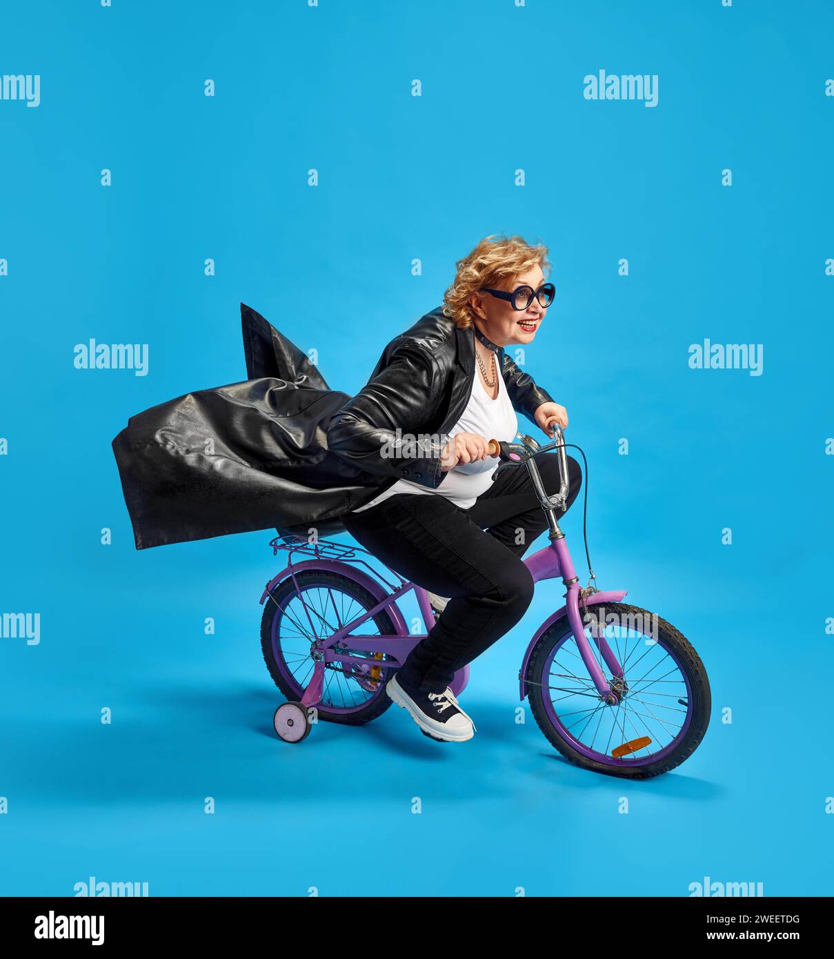 Ältere Frau, die Coolness und Freude ausstrahlt, während sie in Bewegung auf dem lila Kinderfahrrad vor blauem Hintergrund fährt. Stockfoto
