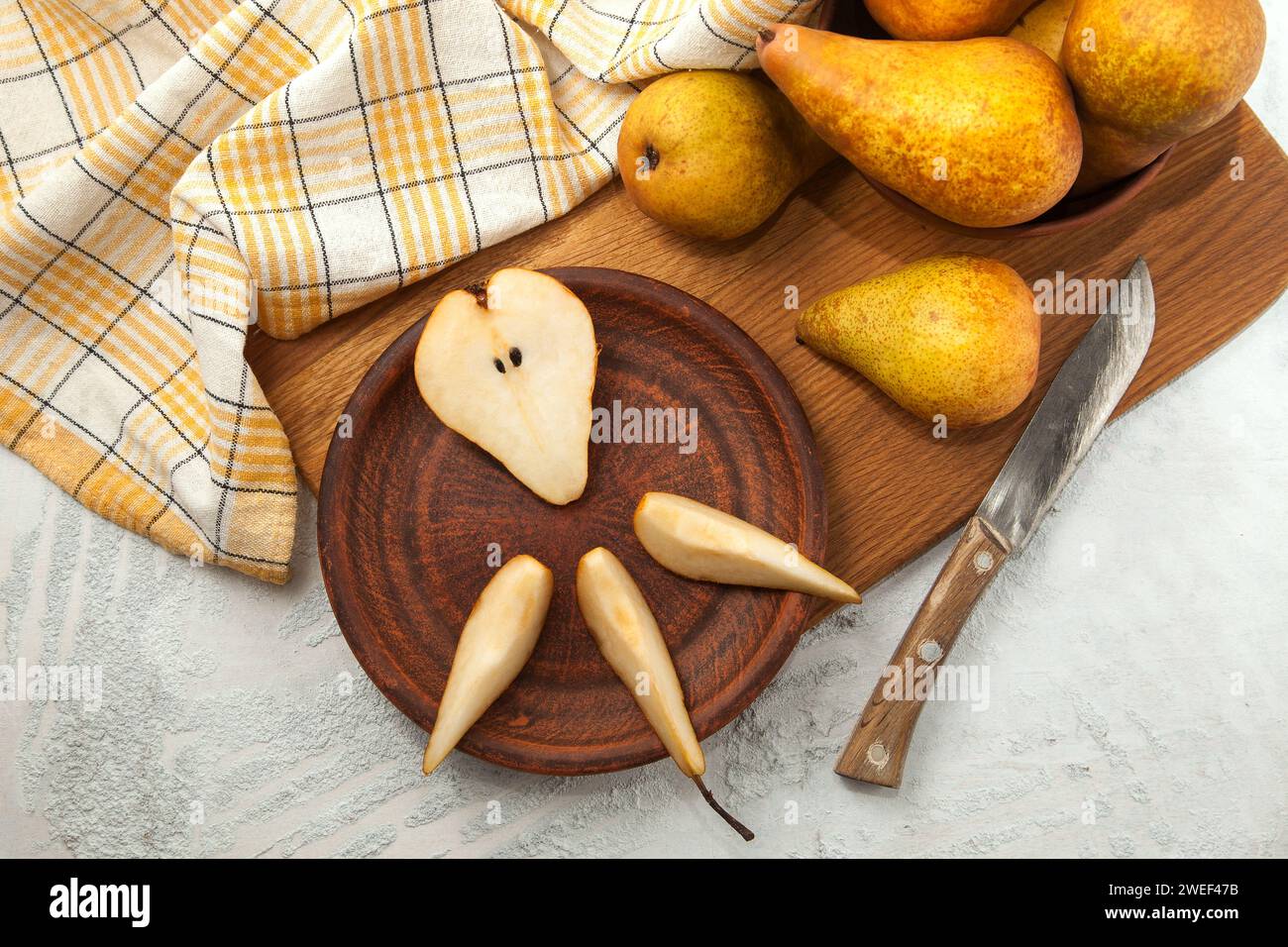 Tonplatte mit Scheiben und der Hälfte reifer saftiger Birnenfrüchte, Korbkorb mit Birnen und gelbes Küchentuch auf Holzschneidebrett. Stockfoto