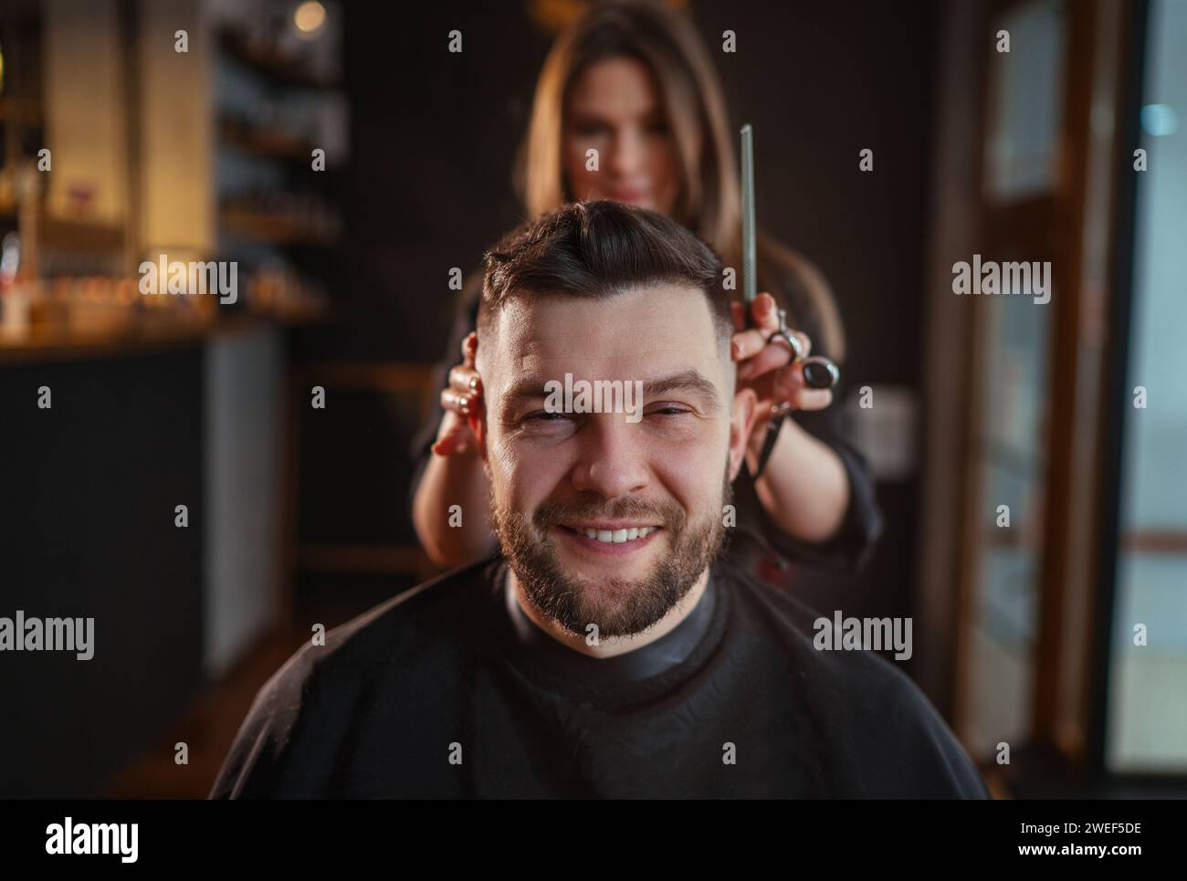 Lächelnder bärtiger Mann Porträt beim Friseurfriseur im Friseursalon von einer jungen Friseurfrau. Moderner Friseurladen in dunklem Schwarz Stockfoto