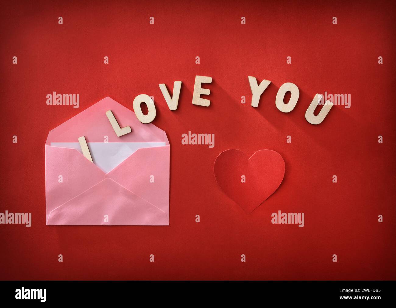 Rosafarbener Umschlag mit romantischem Brief mit Holzbuchstaben mit Nachricht I Love You auf rotem Hintergrund mit Herzausschnitt. Draufsicht. Stockfoto