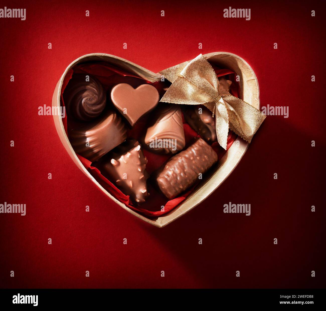 Braune, herzförmige Box gefüllt mit verschiedenen Pralinen mit goldener Schleife auf rotem Hintergrund. Draufsicht. Stockfoto
