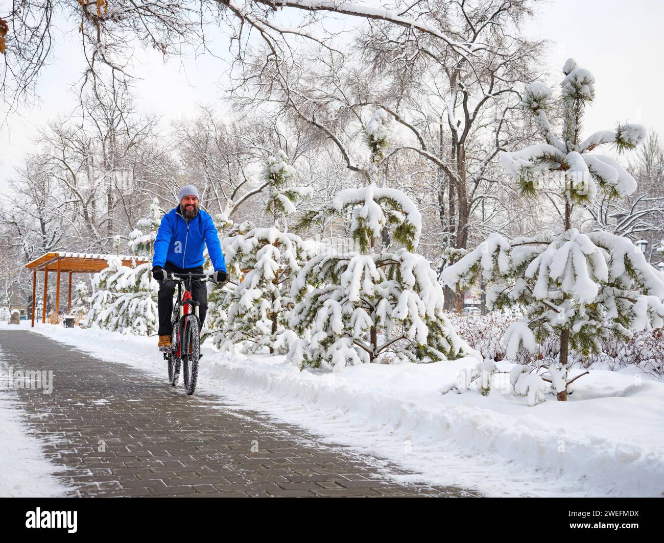 Ein fröhlicher Mann fährt mit dem Fahrrad in einem Winterpark zwischen schneebedeckten Bäumen. Bärtiger Radfahrer in blauer Jacke. Aktiver Lebensstil Stockfoto