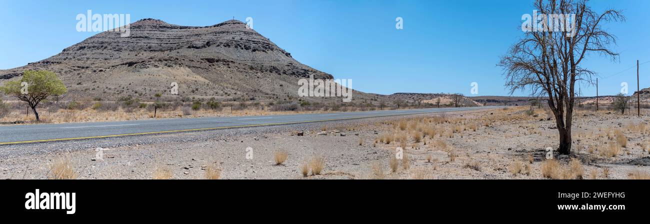 Landschaft mit B4-Teerstraße und geschichteten Basaltfelsen in der Wüste, aufgenommen im hellen Licht des späten Frühlings östlich von Goageb, Namibia, Afrika Stockfoto