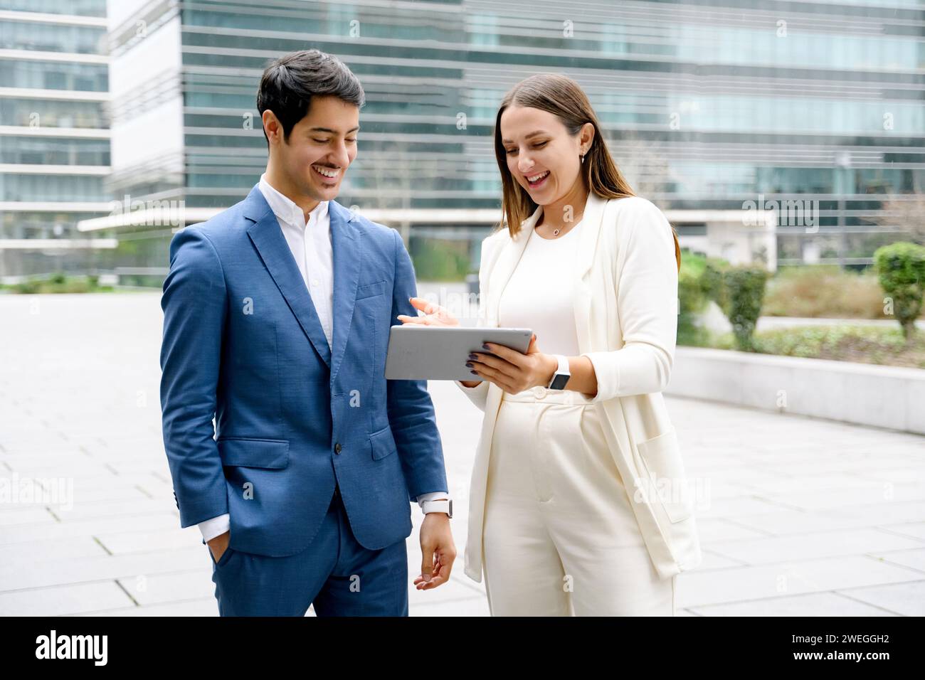 In einem Moment des gemeinsamen Erfolgs sehen ein hispanischer Geschäftsmann und sein Kollege Inhalte auf einem Tablet an, die Teamarbeit und Zusammenarbeit in einer Unternehmensumgebung verkörpern und Geschäftsgebäude im Hintergrund darstellen Stockfoto