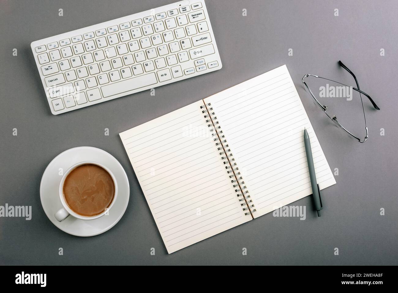 Schreibtisch mit leerem Notizblock, Tastatur, Gläsern und Kaffeetasse auf grauem Tisch. Draufsicht, flach. Stockfoto