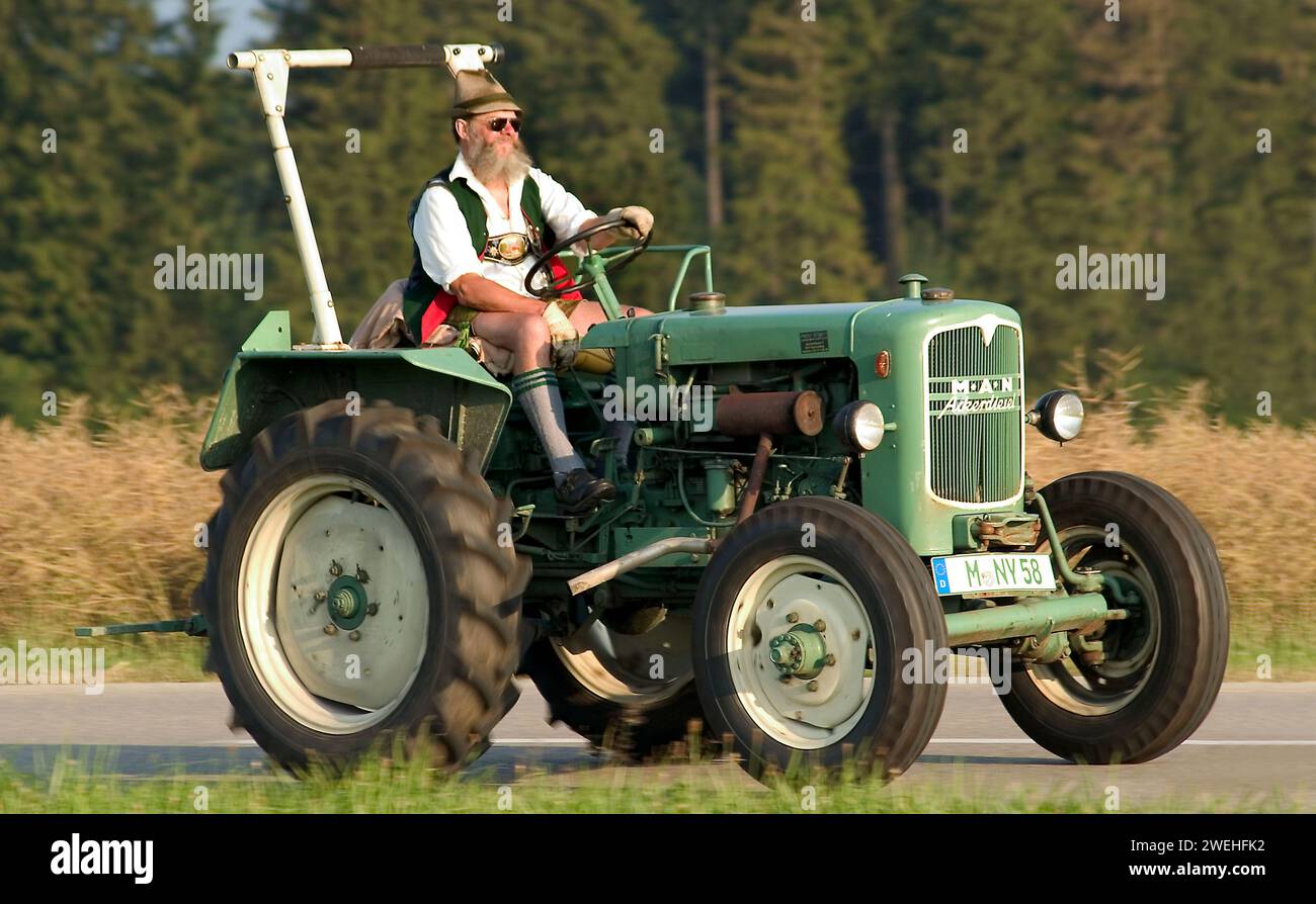 Ein Mann in Lederhosen, mit Vollbart, Hut, Sonnenbrille und traditioneller Tracht sitzt auf einem alten grünen MANN Traktor und fährt durch die Landschaft ne Stockfoto