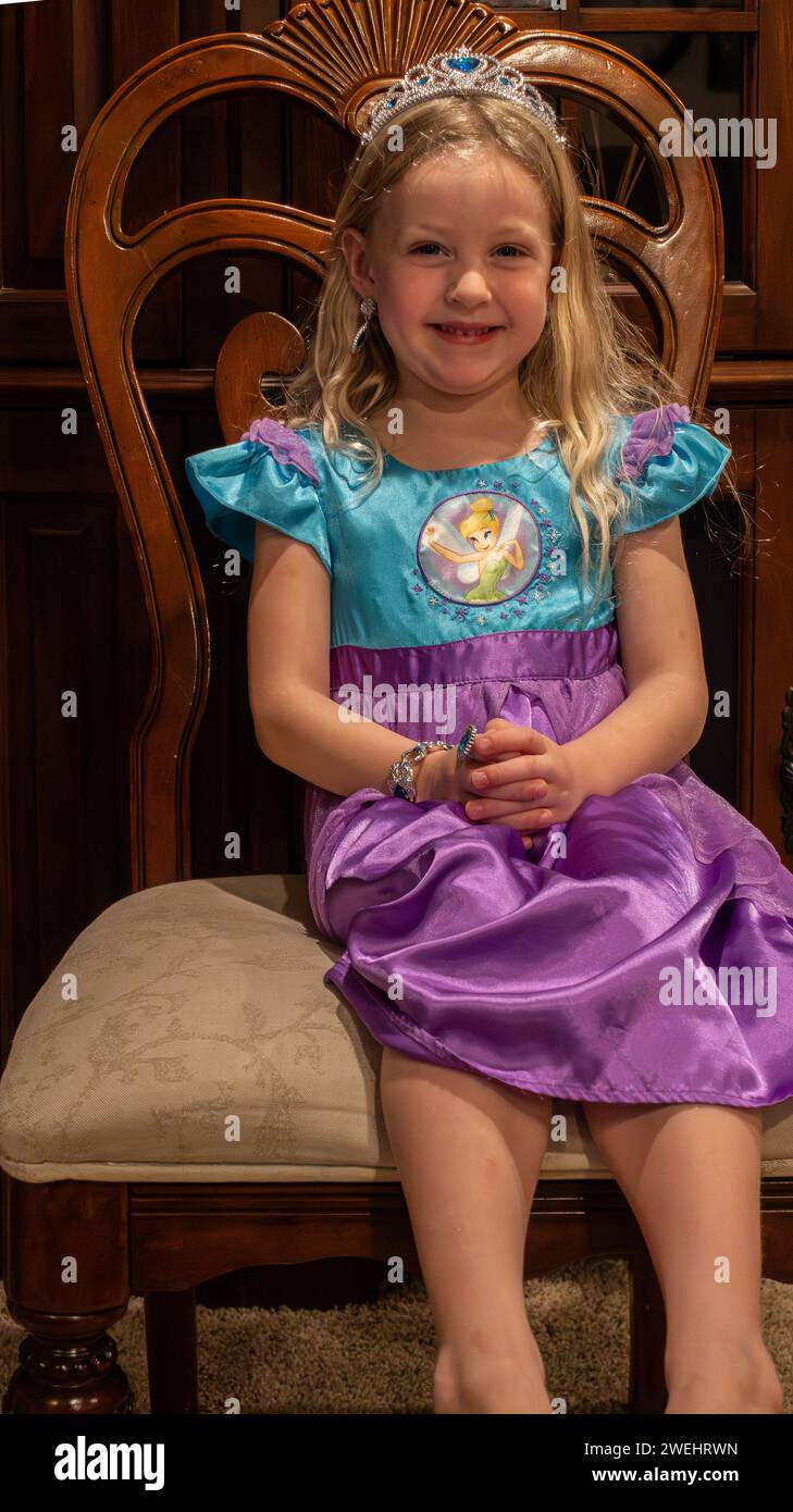 Vier Jahre altes blondes weißes Mädchen, das in einem Stuhl sitzt, während es in einem Disney-Kostüm, Schmuck und Diadem gekleidet ist, während es Rollenspiele spielt. Stockfoto