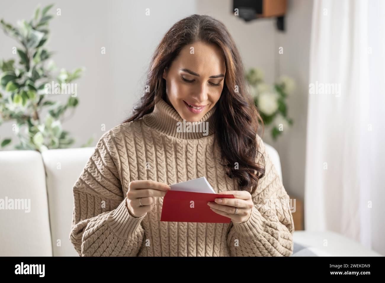Eine gut aussehende Brünette liest einen Liebesbrief mit einem roten Umschlag, den sie von ihrem Freund erhalten hat. Stockfoto