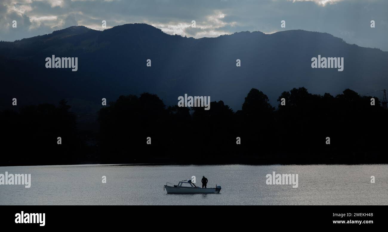 Eine Einzelperson auf einem kleinen Boot, die durch das ruhige Wasser eines Sees gegen eine Bergkette navigiert Stockfoto