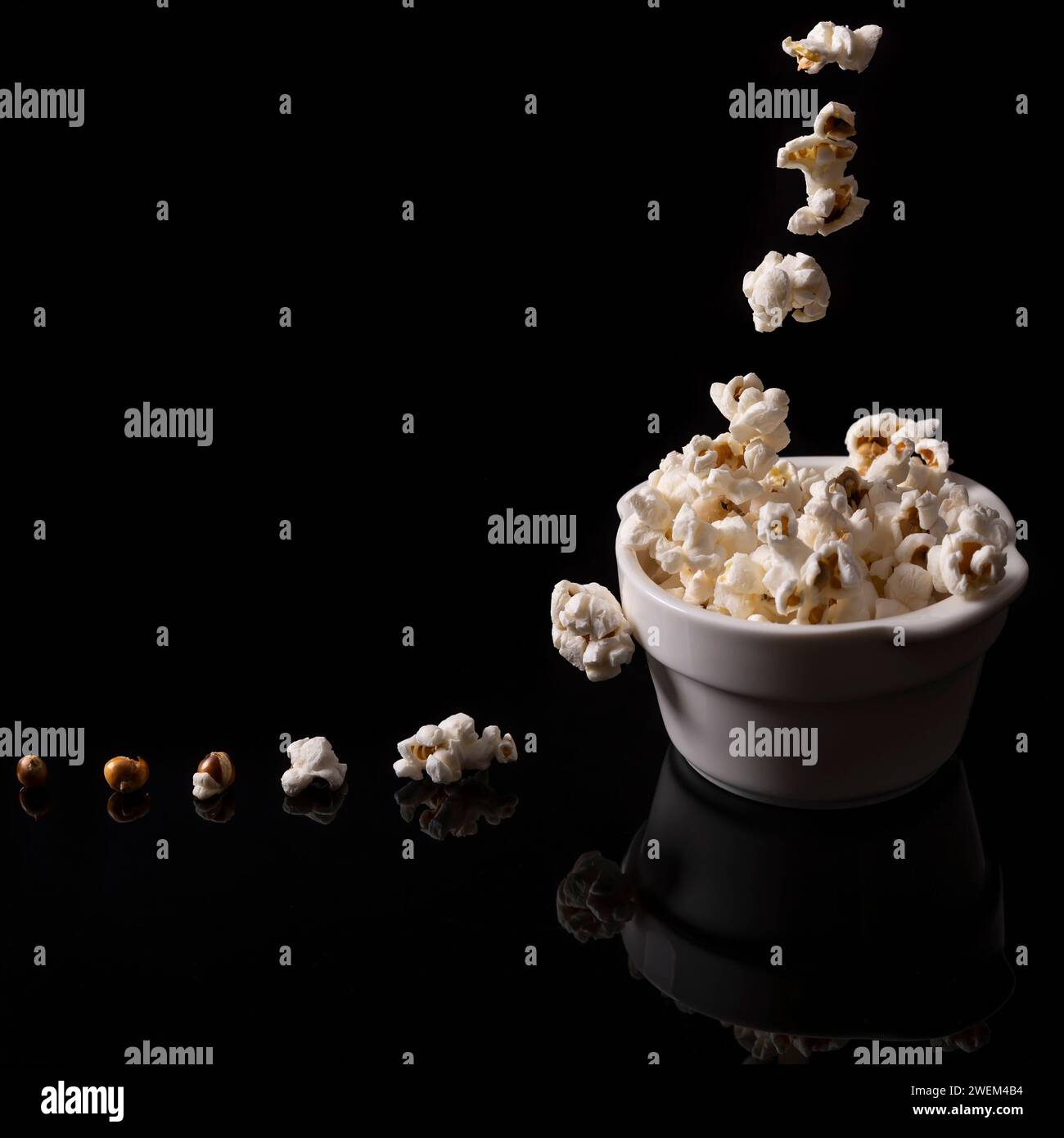 Popcorn fällt in eine weiße Keramikschale und auf dem glänzend schwarzen Hintergrund mit Reflexion Stockfoto