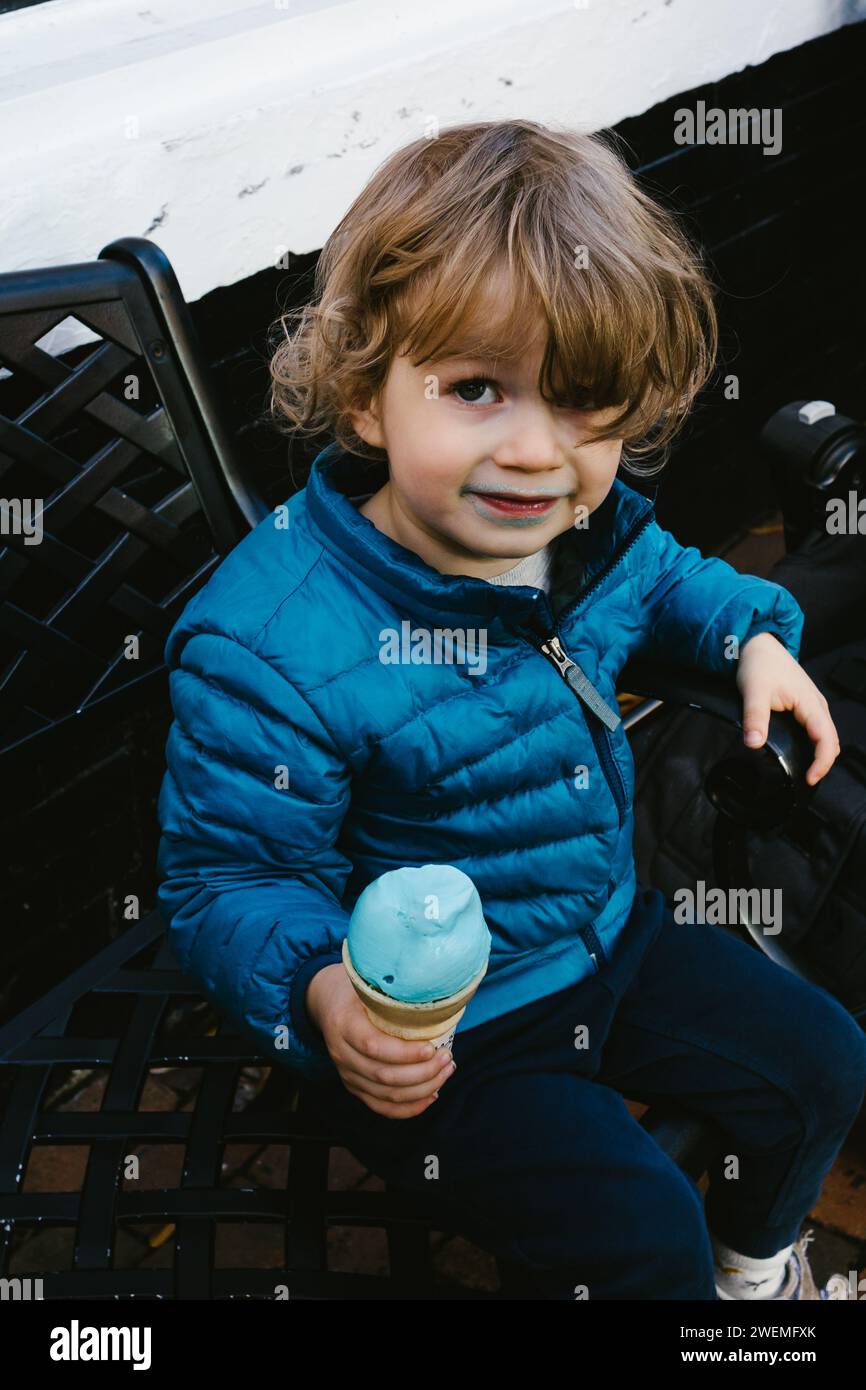 Kleiner Junge im blauen Mantel isst blaues Eis auf der Bank der Stadt Stockfoto