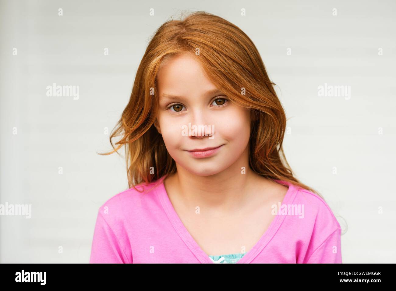 Außenporträt eines süßen kleinen 8-9-jährigen Mädchens mit langen roten Haaren, in rosa Jacke, stehend vor weißem Hintergrund Stockfoto